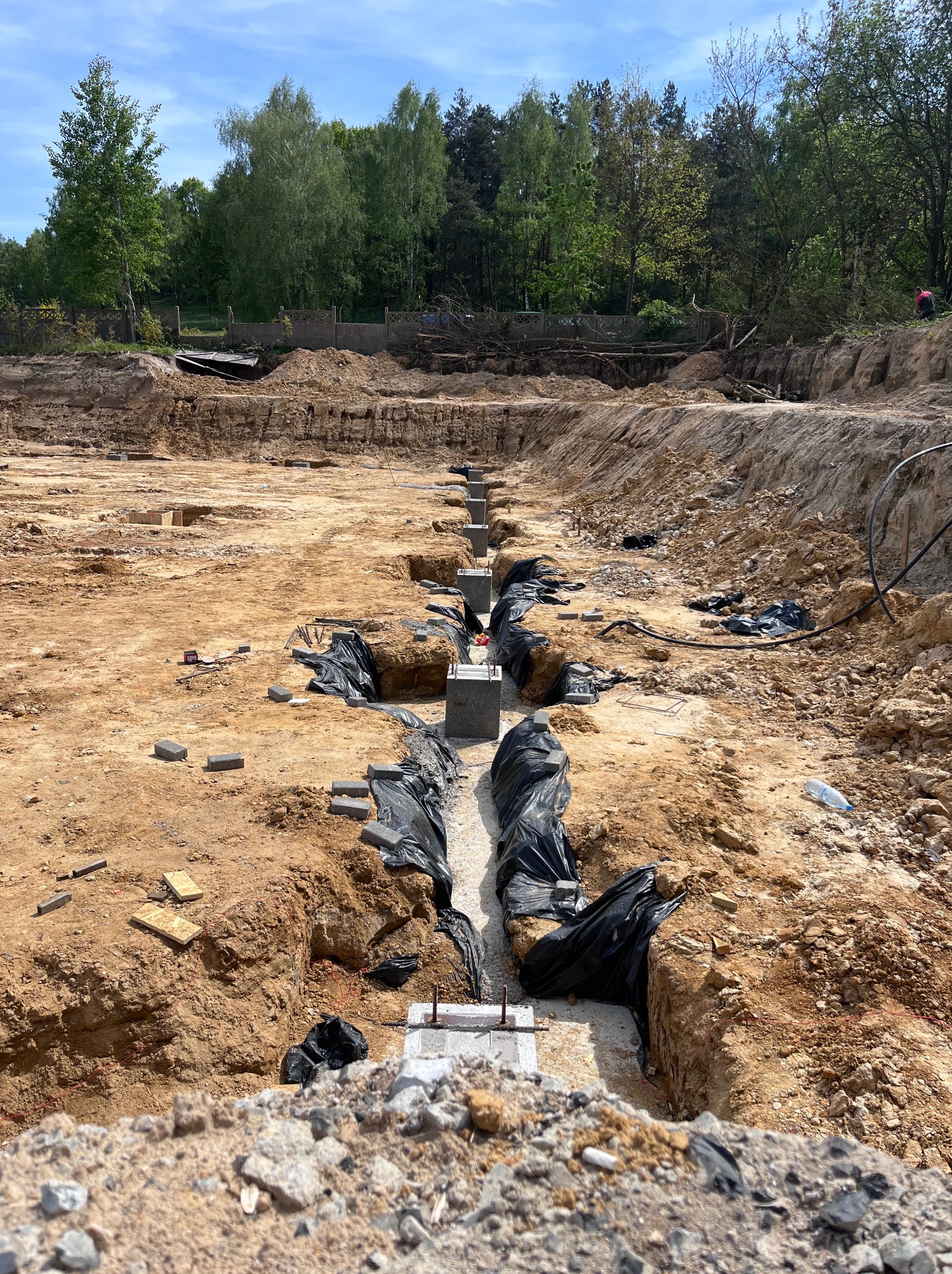 Wykonane fundamenty pod halę w Okmianach, widoczne betonowe słupy i czarna folia ochronna w wykopach, w tle drzewa i niebieskie niebo.