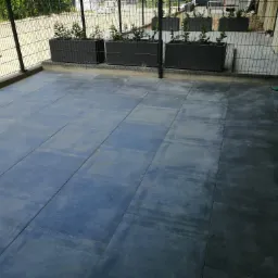 Wyłożony grafitowymi płytkami taras z metalową balustradą i donicami z zielonymi roślinami, widok na zaparkowany samochód