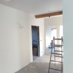 ProBud - Wnętrze w trakcie odświeżania z pomalowanymi na biało ścianami, drewnianą belką sufitową z wiszącą żarówką, widoczną drabiną i surową podłogą z plamami farby.