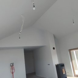 ProBud - Surowe wnętrze w Kamieniu Pomorskim po szpachlowaniu i malowaniu, widoczne przygotowane do montażu oświetlenie, grzejnik typu koza i lampa budowlana.