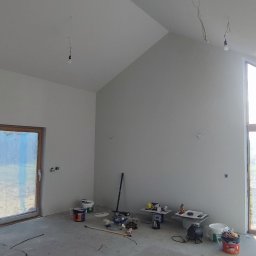 ProBud - Surowe wnętrze z wysokim sufitem po malowaniu na szaro, widoczne okno z folią ochronną, narzędzia malarskie, niedokończone gniazdka elektryczne i żarówki na kablach.