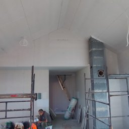 ProBud - Wnętrze w trakcie prac wykończeniowych: ściany i sufit obłożone płytami gipsowo-kartonowymi, widoczne rusztowania, przewody elektryczne zwisające z sufitu, fragment komina z szarego bloczka...