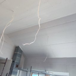 ProBud - Wnętrze w trakcie wykończenia: ściany i sufit pokryte płytami gipsowo-kartonowymi, widoczne przewody elektryczne zwisające z sufitu, fragment rusztowania i komina.
