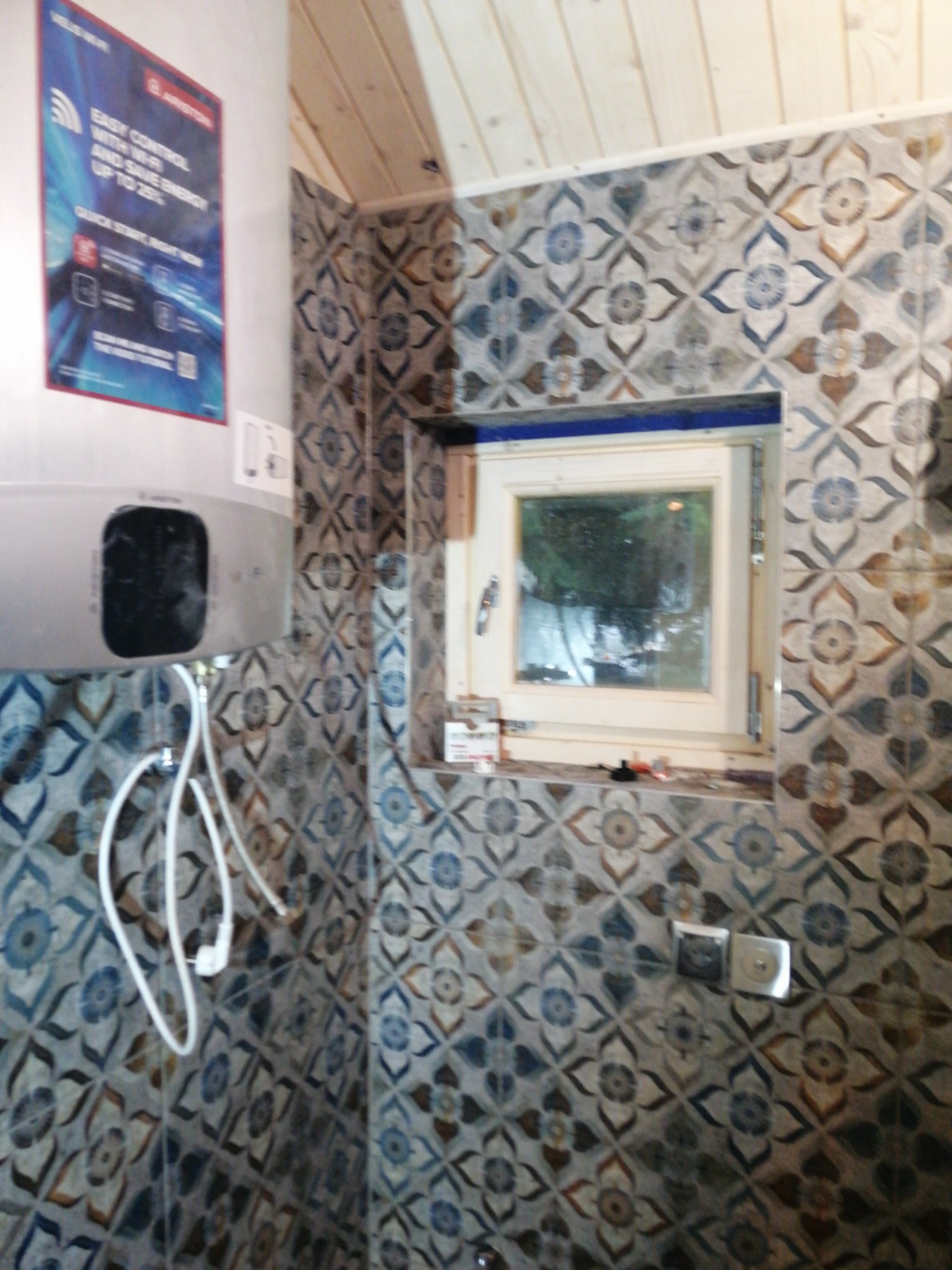 Wnętrze łazienki z wzorzystymi, niebiesko-brązowymi płytkami, widoczny bojler elektryczny z reklamą producenta, okno z drewnianą ramą oraz drewniana boazeria na suficie.