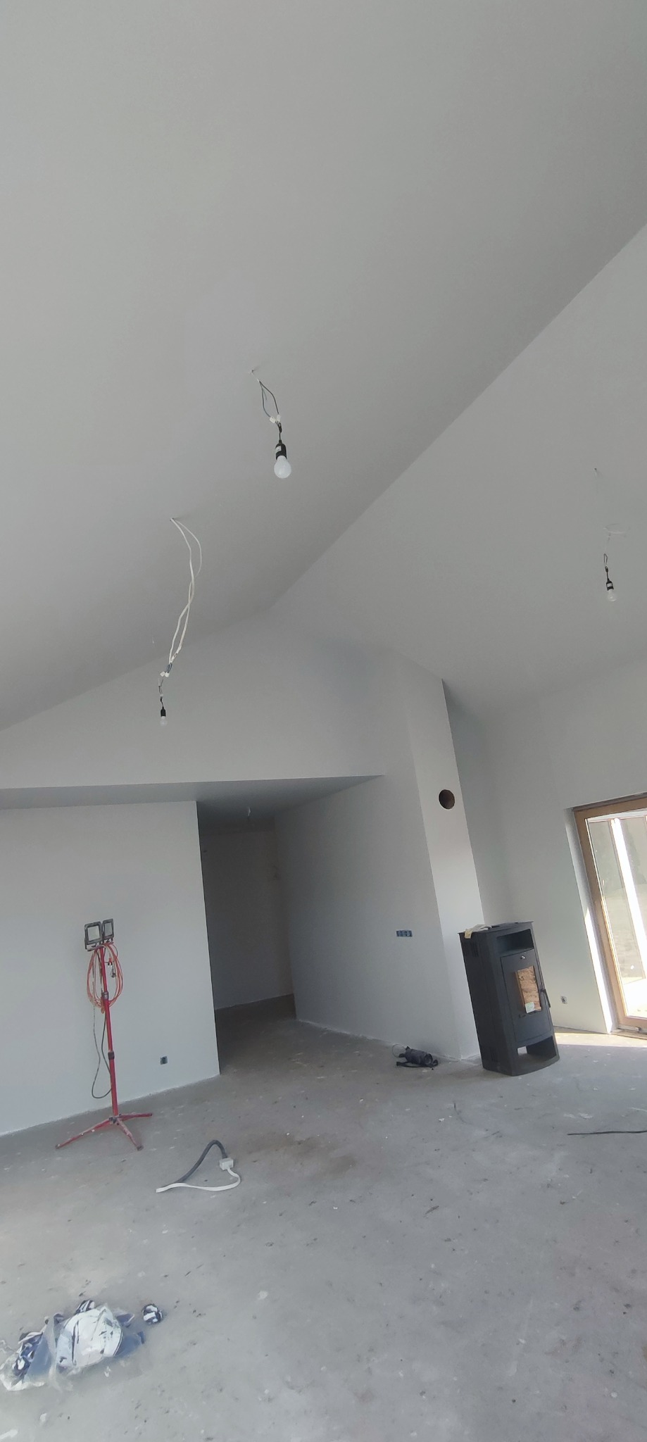 Surowe wnętrze po szpachlowaniu i malowaniu na biało, widoczne wiszące kable, lampa budowlana, piec wolnostojący i okno. Podłoga betonowa.
