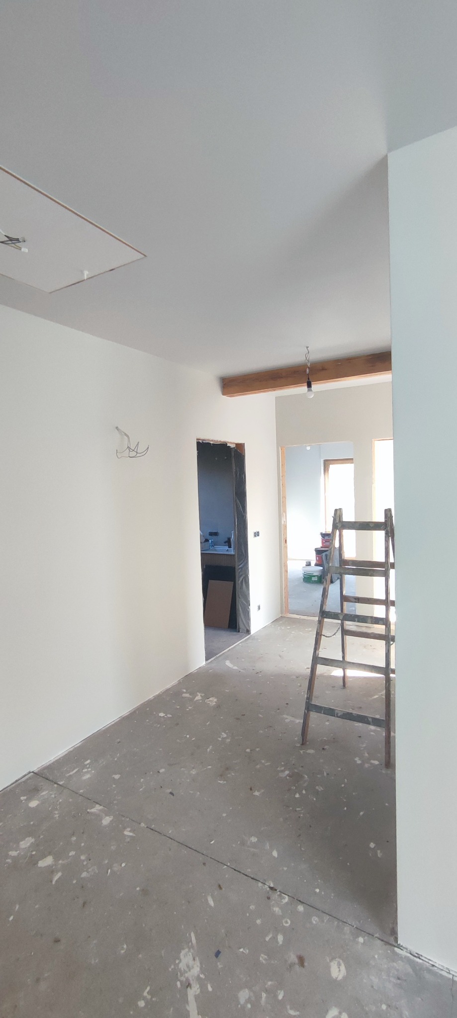 Wnętrze w trakcie odświeżania z pomalowanymi na biało ścianami, drewnianą belką sufitową z wiszącą żarówką, widoczną drabiną i surową podłogą z plamami farby.