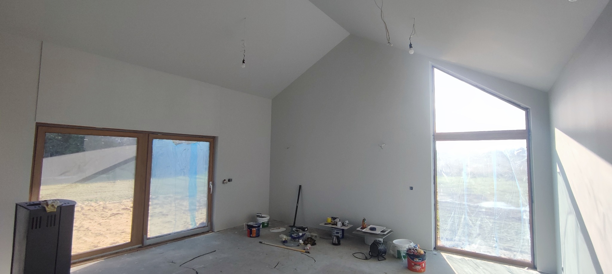 Surowe wnętrze z wysokim sufitem po malowaniu na szaro, widoczne okno z folią ochronną, narzędzia malarskie, niedokończone gniazdka elektryczne i żarówki na kablach.