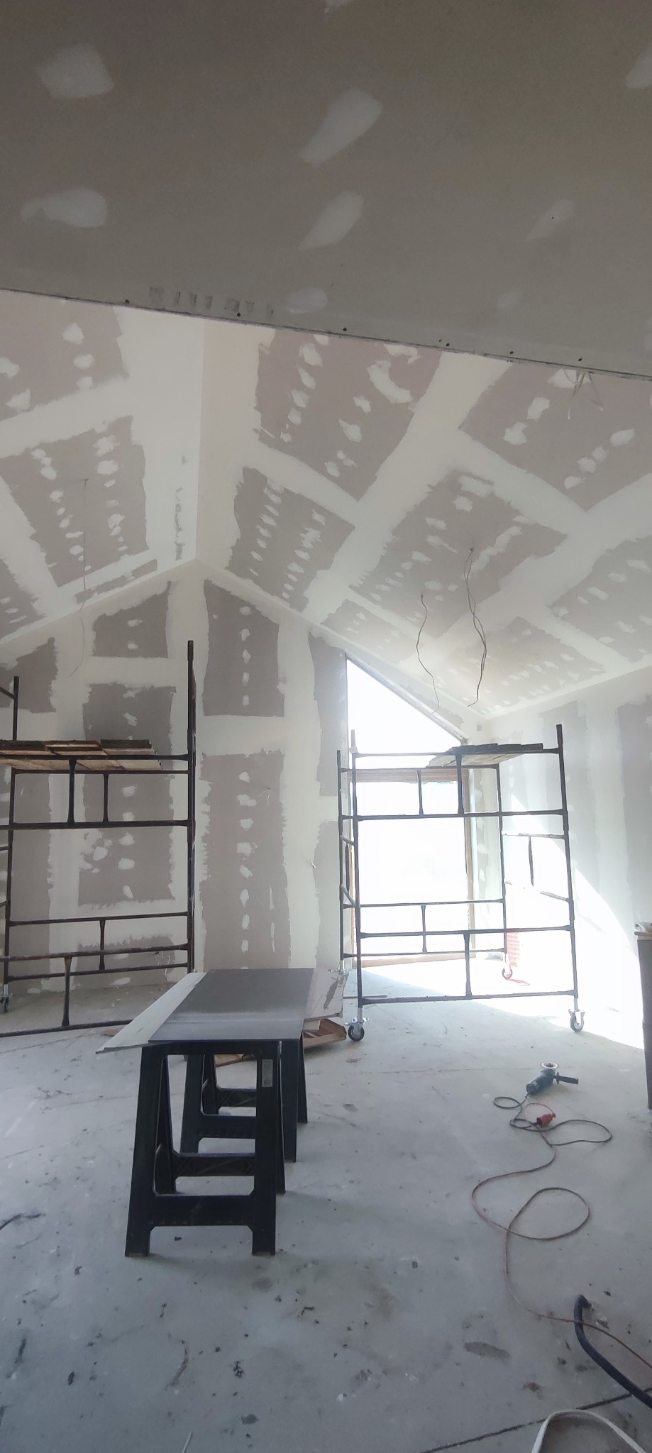 Skośny sufit i ściany z płyt gipsowo-kartonowych po szpachlowaniu, widoczne rusztowania, szlifierka kątowa na podłodze i stół roboczy w pomieszczeniu z oknem.