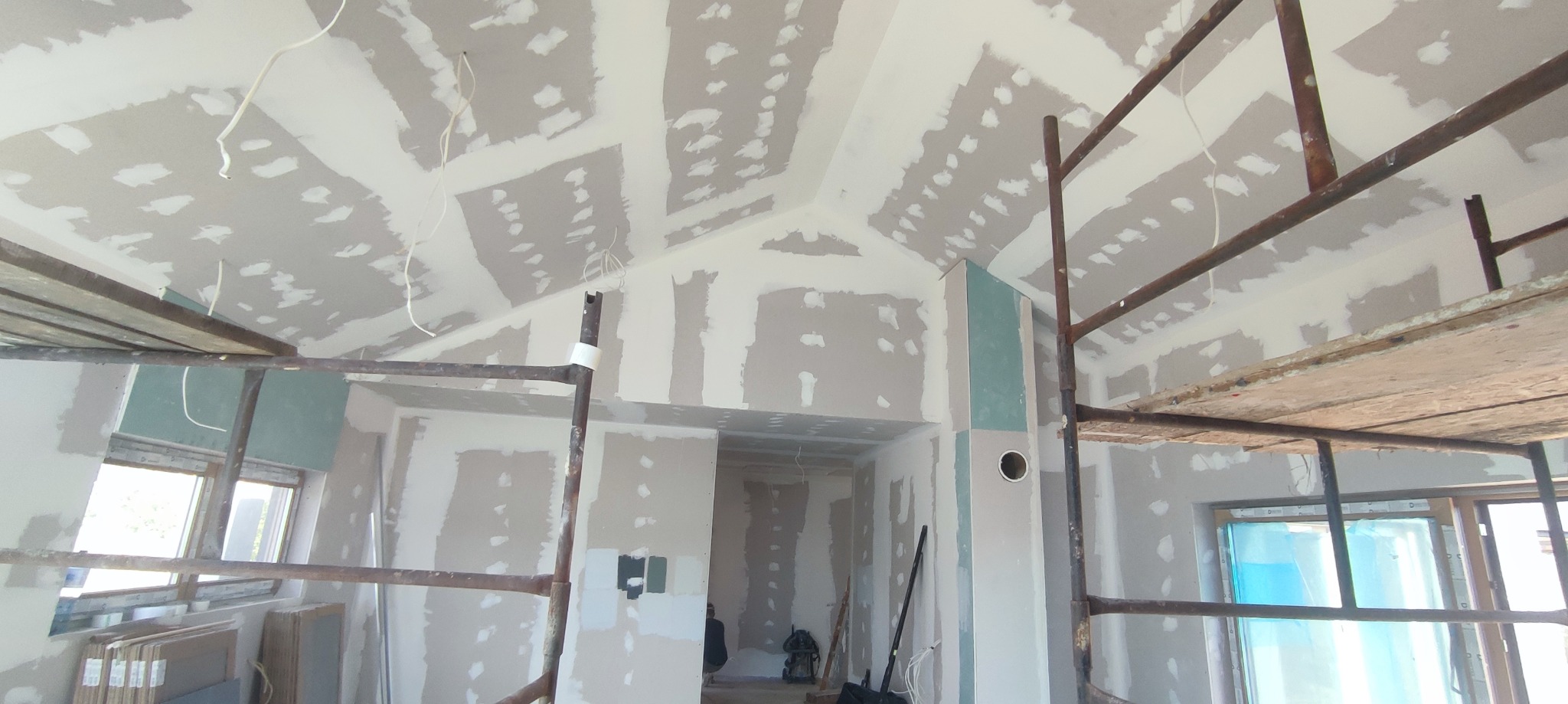 Sufit i ściany w trakcie szpachlowania, widoczne rusztowania, instalacja elektryczna wystająca z sufitu, surowe wnętrze w trakcie prac wykończeniowych.