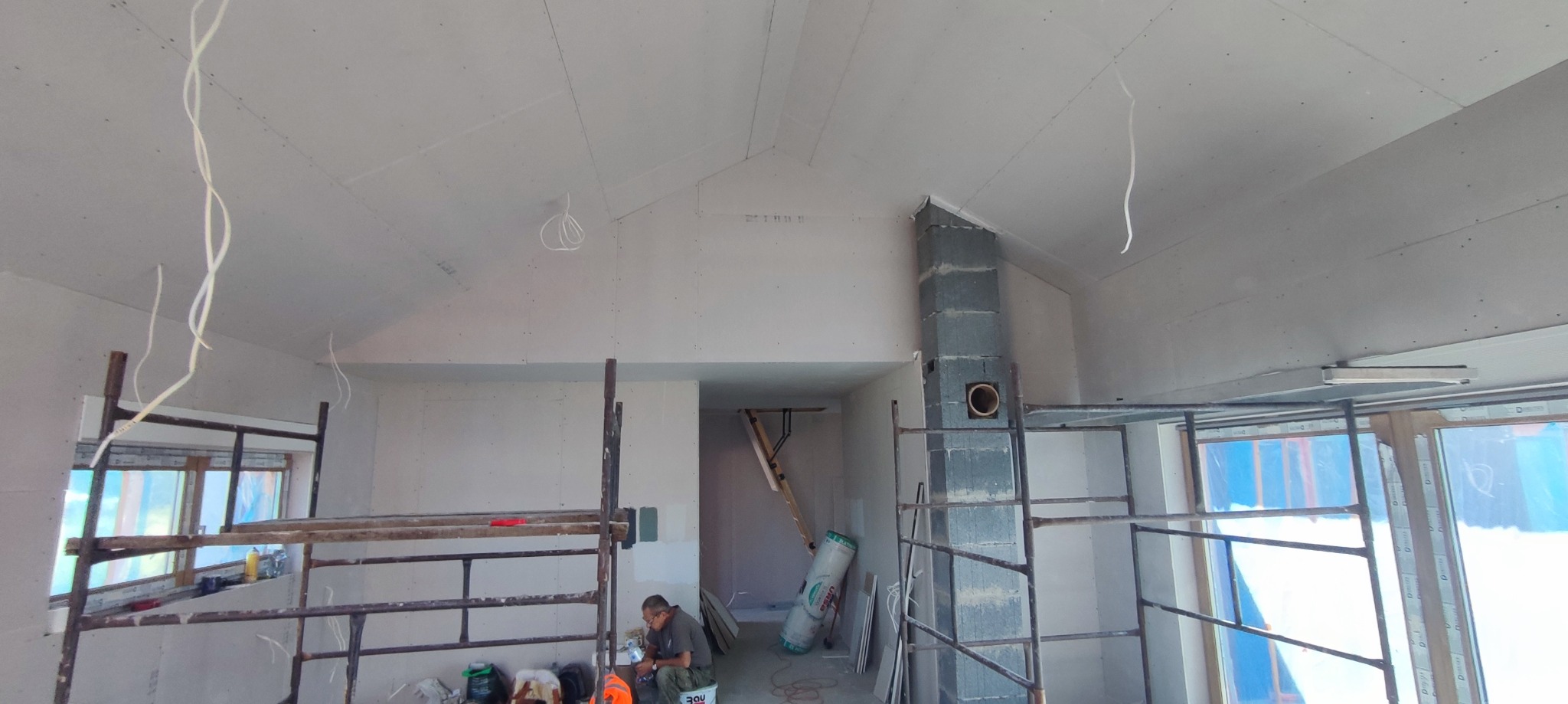 Wnętrze w trakcie prac wykończeniowych: ściany i sufit obłożone płytami gipsowo-kartonowymi, widoczne rusztowania, przewody elektryczne zwisające z sufitu, fragment komina z szarego bloczka...