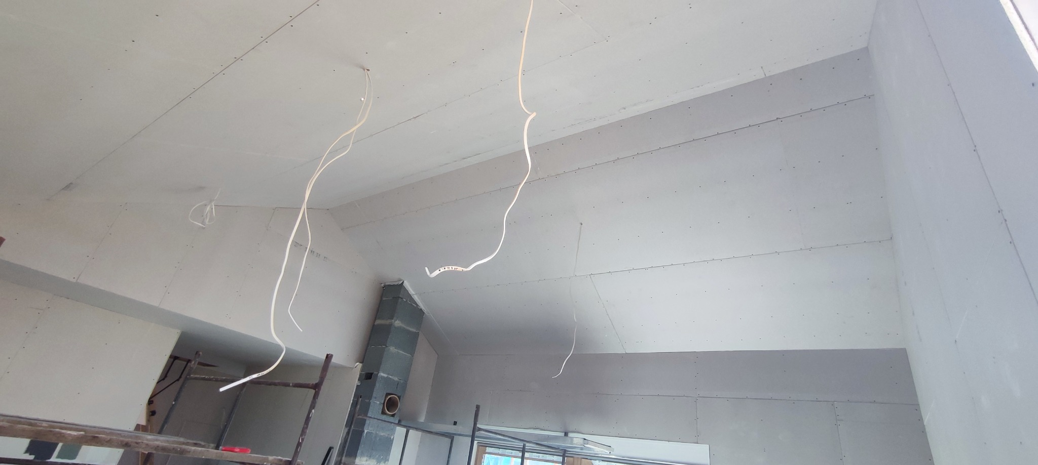 Wnętrze w trakcie wykończenia: ściany i sufit pokryte płytami gipsowo-kartonowymi, widoczne przewody elektryczne zwisające z sufitu, fragment rusztowania i komina.