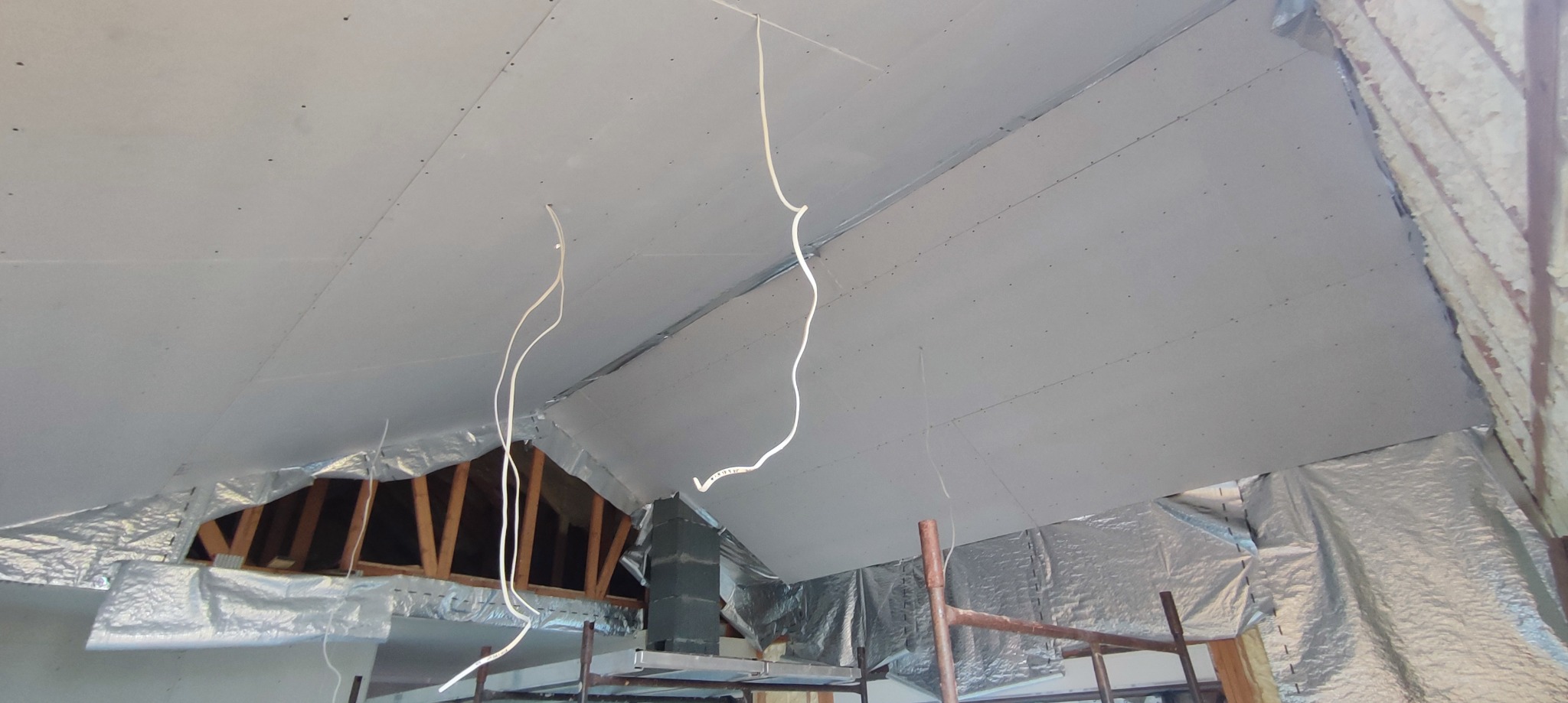 Montaż płyt gipsowo-kartonowych na poddaszu, widoczne kable zwisające z sufitu i fragment izolacji termicznej, rusztowanie pomocnicze w tle.
