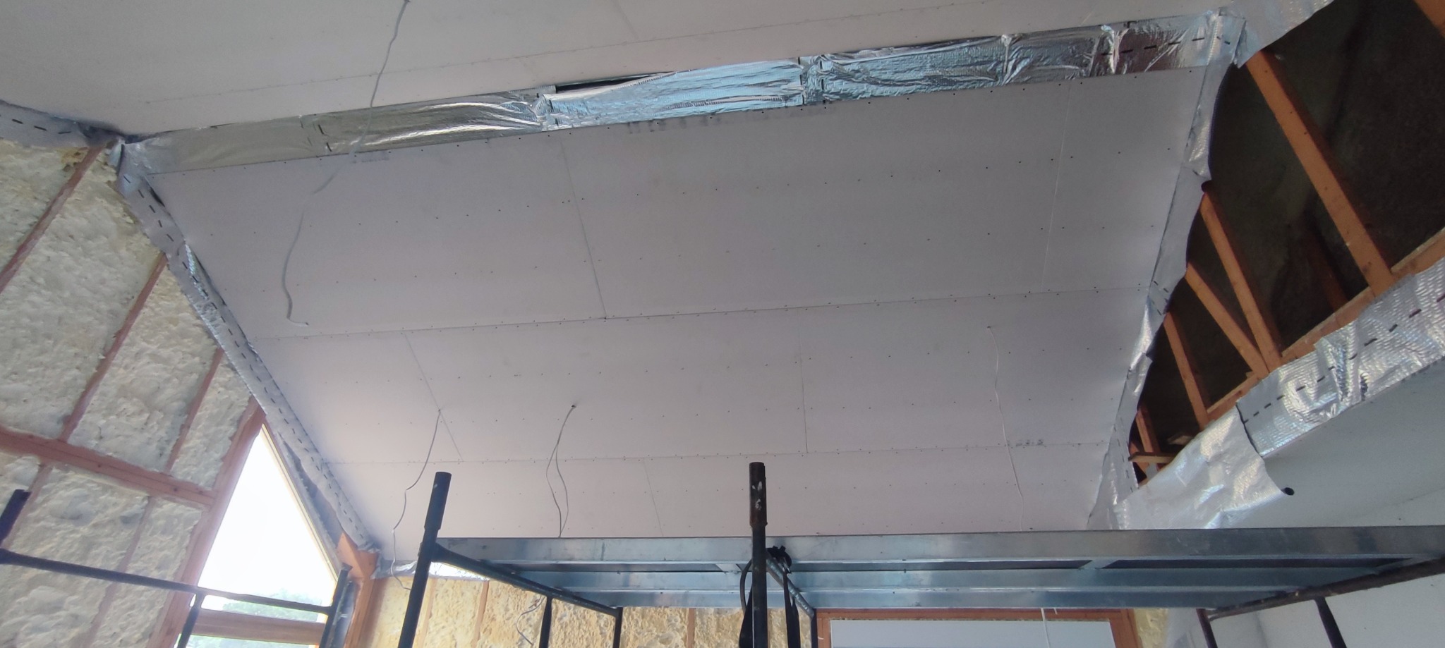 Montaż płyt gipsowo-kartonowych na skośnym poddaszu, widoczne izolacja folią aluminiową, rusztowanie i drewniana konstrukcja dachu