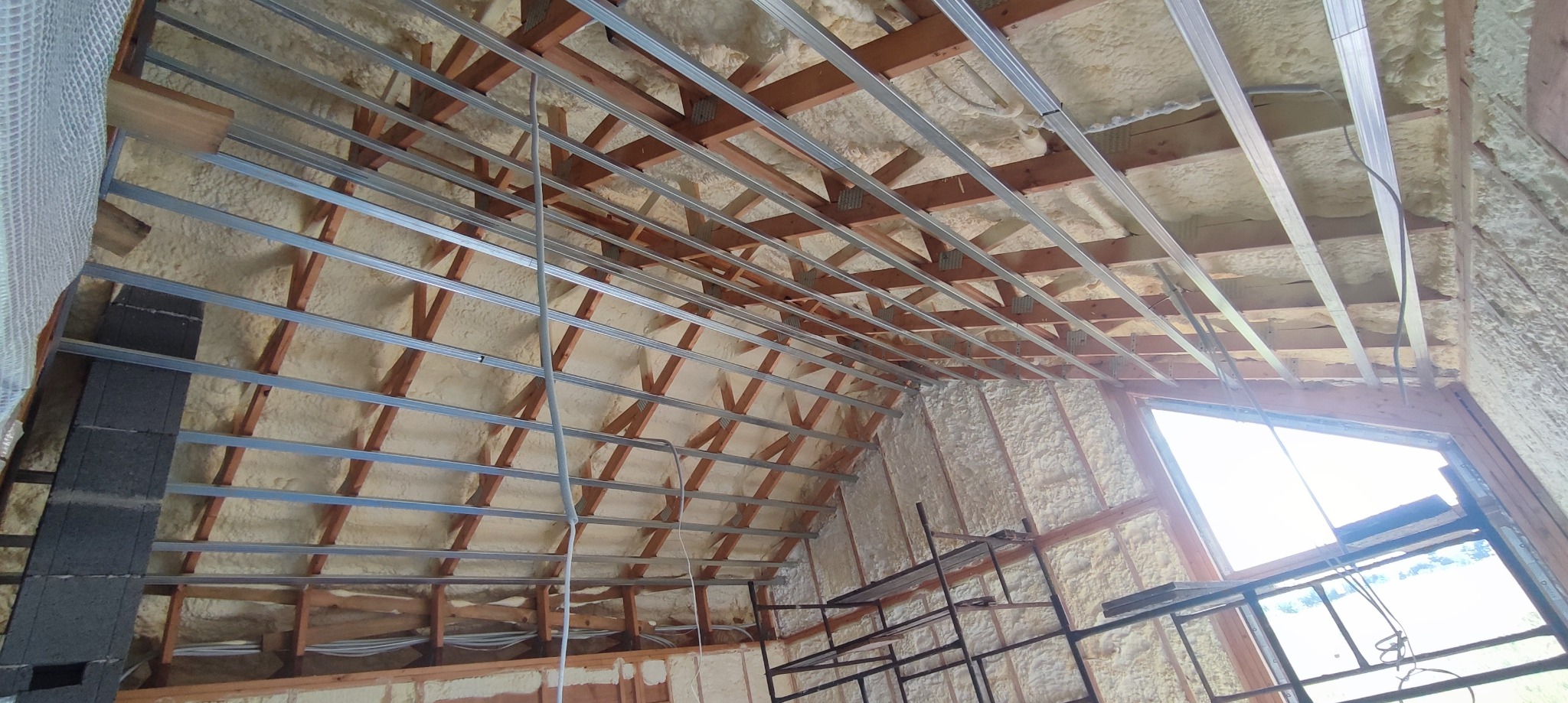 Poddasze ocieplone pianą PUR z widoczną drewnianą konstrukcją dachu i metalowymi profilami pod płyty gipsowo-kartonowe, okno dachowe z widokiem na zewnątrz.