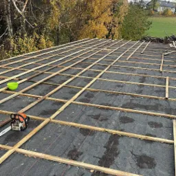 Montaż drewnianej konstrukcji dachu pokrytego papą, widoczna piła spalinowa Stihl i miska, drzewa w tle.