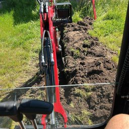 Mont Bud Team Jakub Trzciński - Czerwona mini koparka z perspektywy operatora, wykonująca wykop w gruncie. Widoczny budynek w tle i poziomica oparta o ścianę.