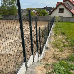 Nowoczesne ogrodzenie panelowe z podmurówką betonową, widok z boku na tle domów jednorodzinnych i zieleni. Zastosowano metalowe słupki i siatkę w kolorze grafitowym.