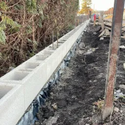 Wznoszenie fundamentu pod ogrodzenie z bloczków betonowych. Widoczne zbrojenie i folia izolacyjna. Tło: tuje i drzewa. Perspektywa z dołu, podkreślająca długość fundamentu.