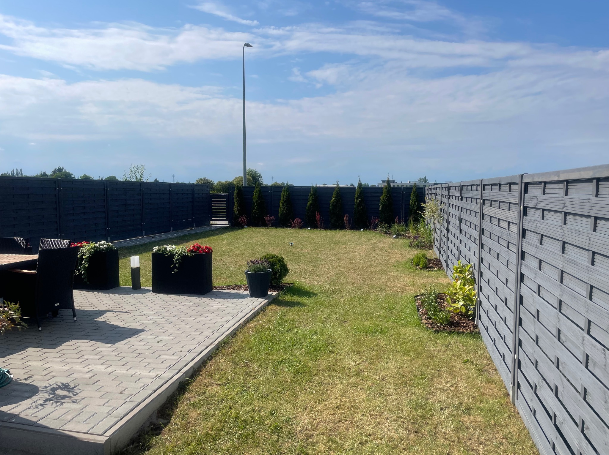 Widok na zadbany ogród z drewnianym ogrodzeniem, trawnikiem, tujami, kostką brukową na tarasie i dekoracyjnymi donicami z kwiatami w słoneczny dzień.