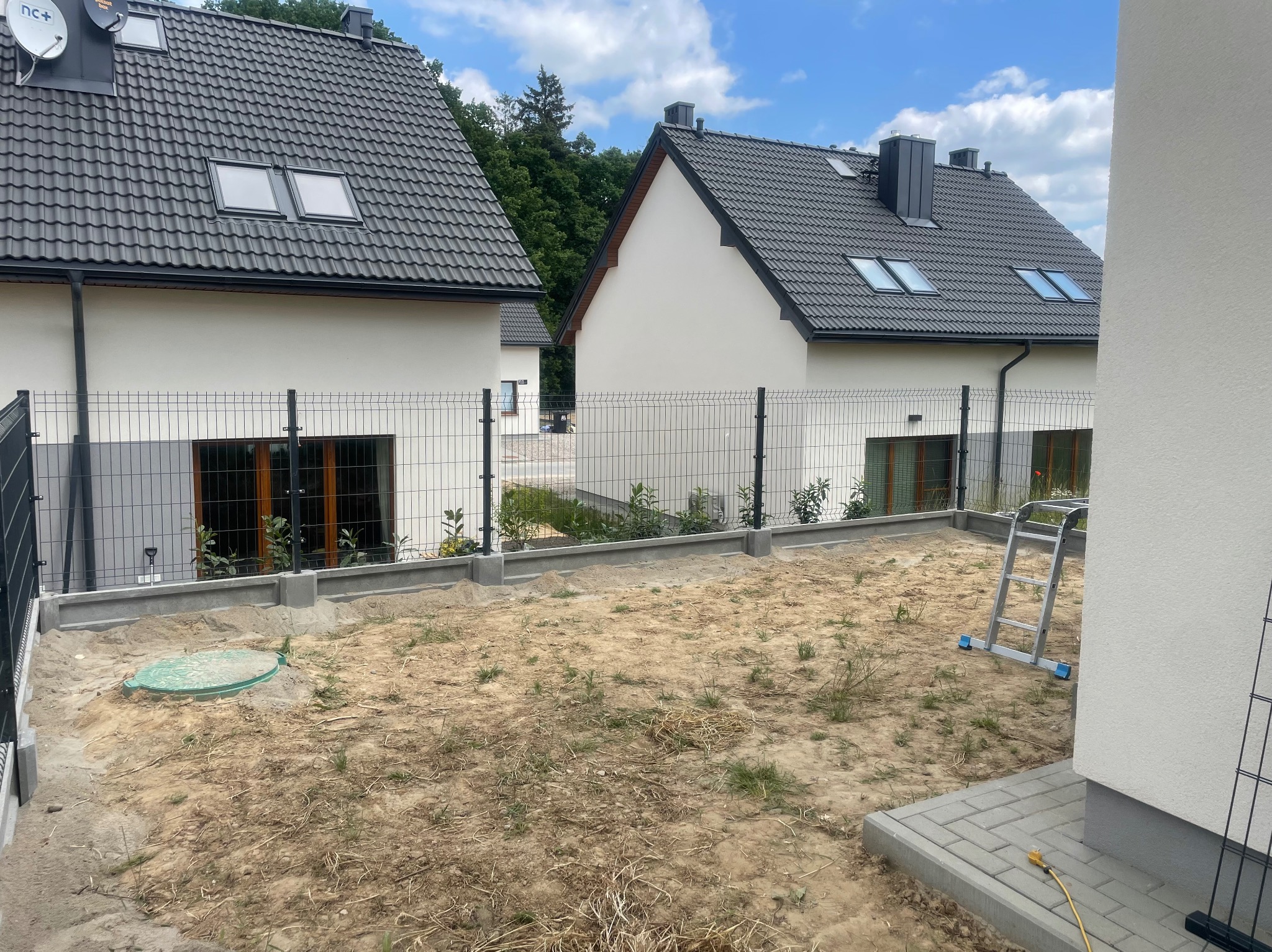 Teren działki z dwoma nowymi domami w tle, ogrodzony siatką, z widoczną studzienką kanalizacyjną i drabiną opartą o ścianę, przygotowany pod przyszły ogród.