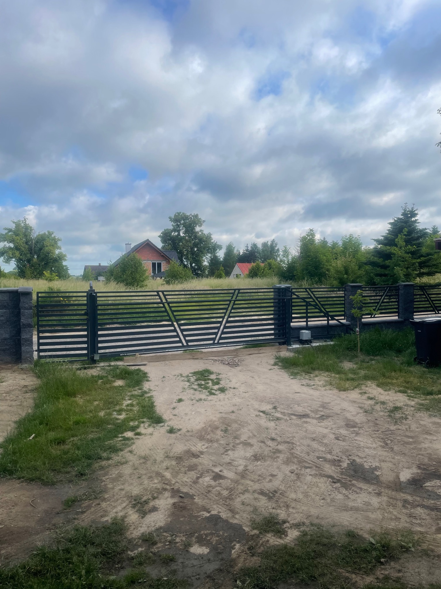 Nowoczesna, pozioma brama wjazdowa z szarymi słupkami z imitacji kamienia prowadząca do posesji z domem w tle, widok w pochmurny dzień.