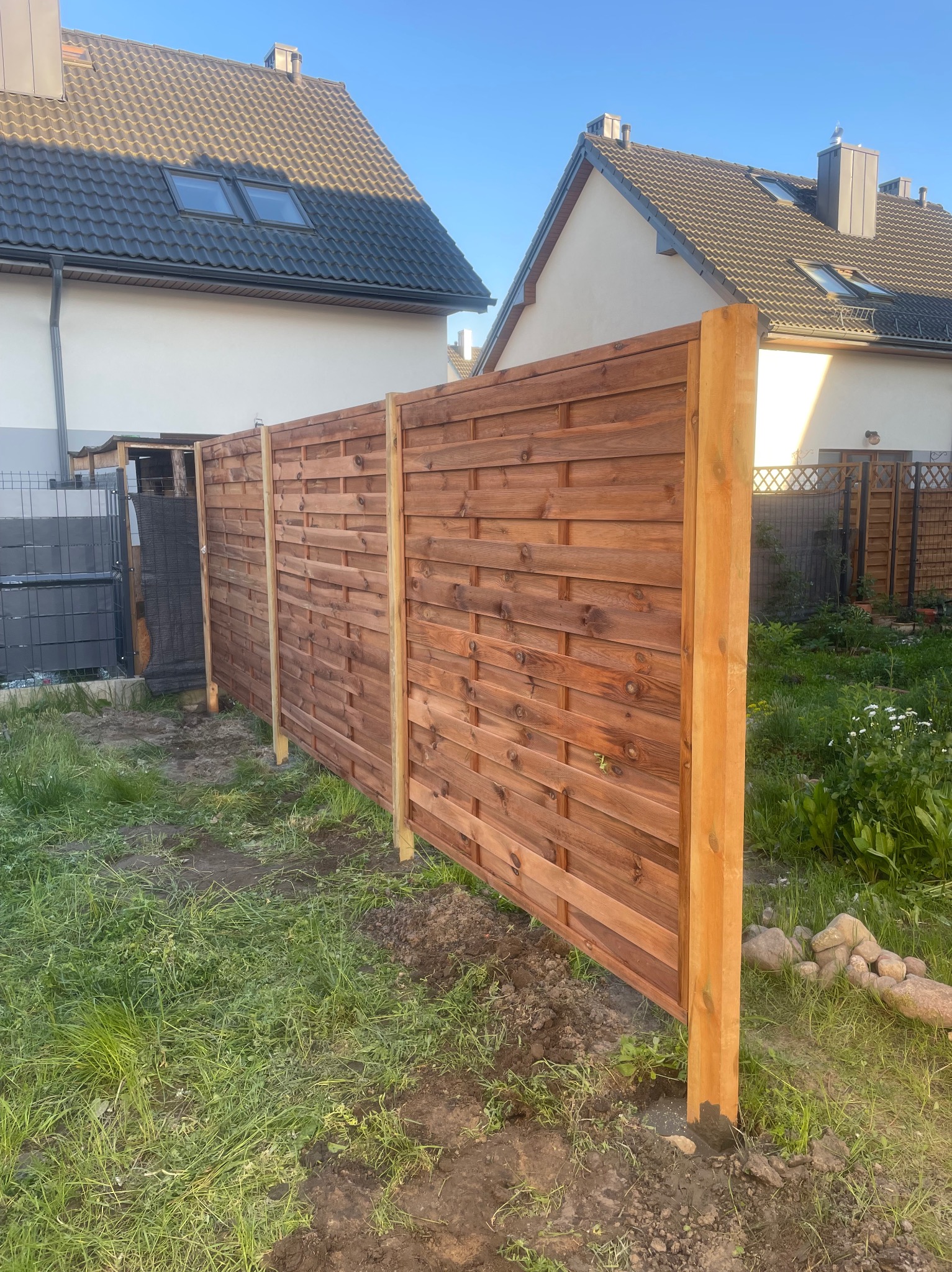 Nowo postawiony, drewniany płot panelowy o poziomej strukturze desek, z widocznymi słupkami, na tle domów jednorodzinnych i zielonego trawnika.