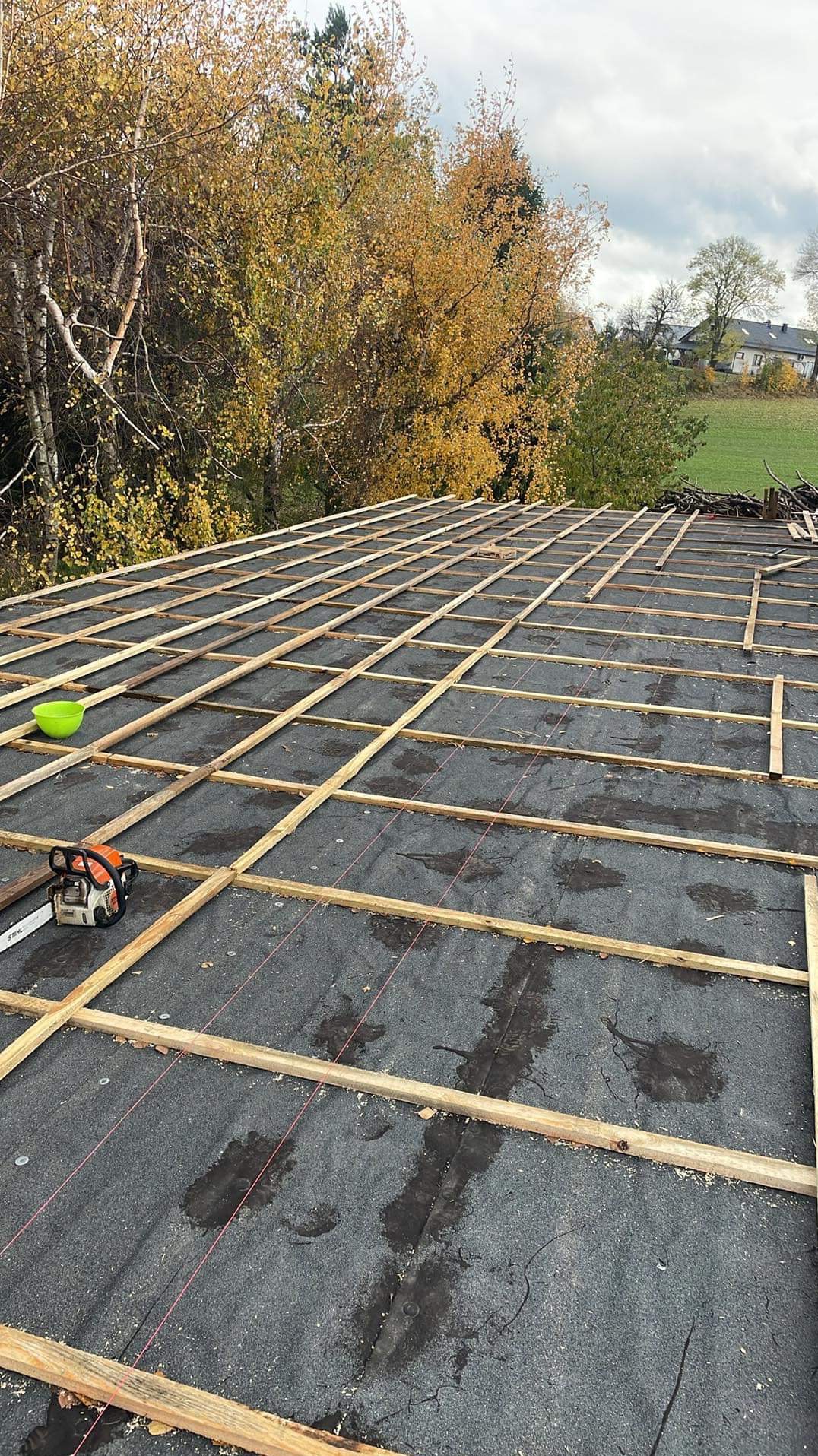 Montaż drewnianej konstrukcji dachu pokrytego papą, widoczna piła spalinowa Stihl i miska, drzewa w tle.