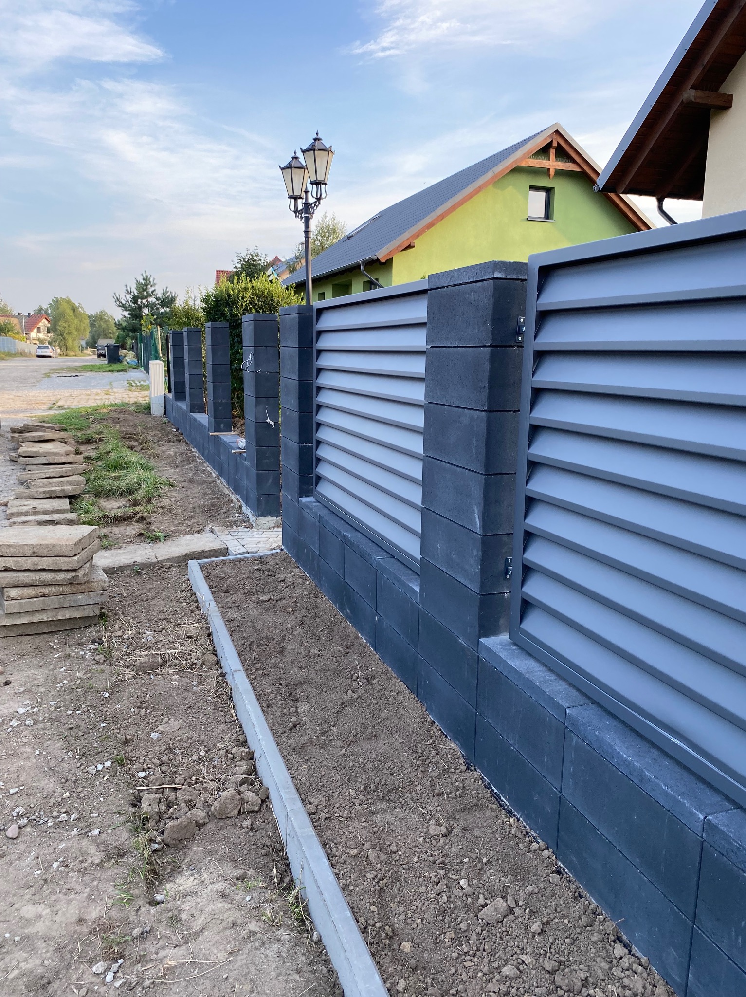 Montaż nowoczesnego ogrodzenia panelowego z betonowych bloczków w kolorze grafitowym, widoczne przygotowane podłoże i ułożone płyty chodnikowe.