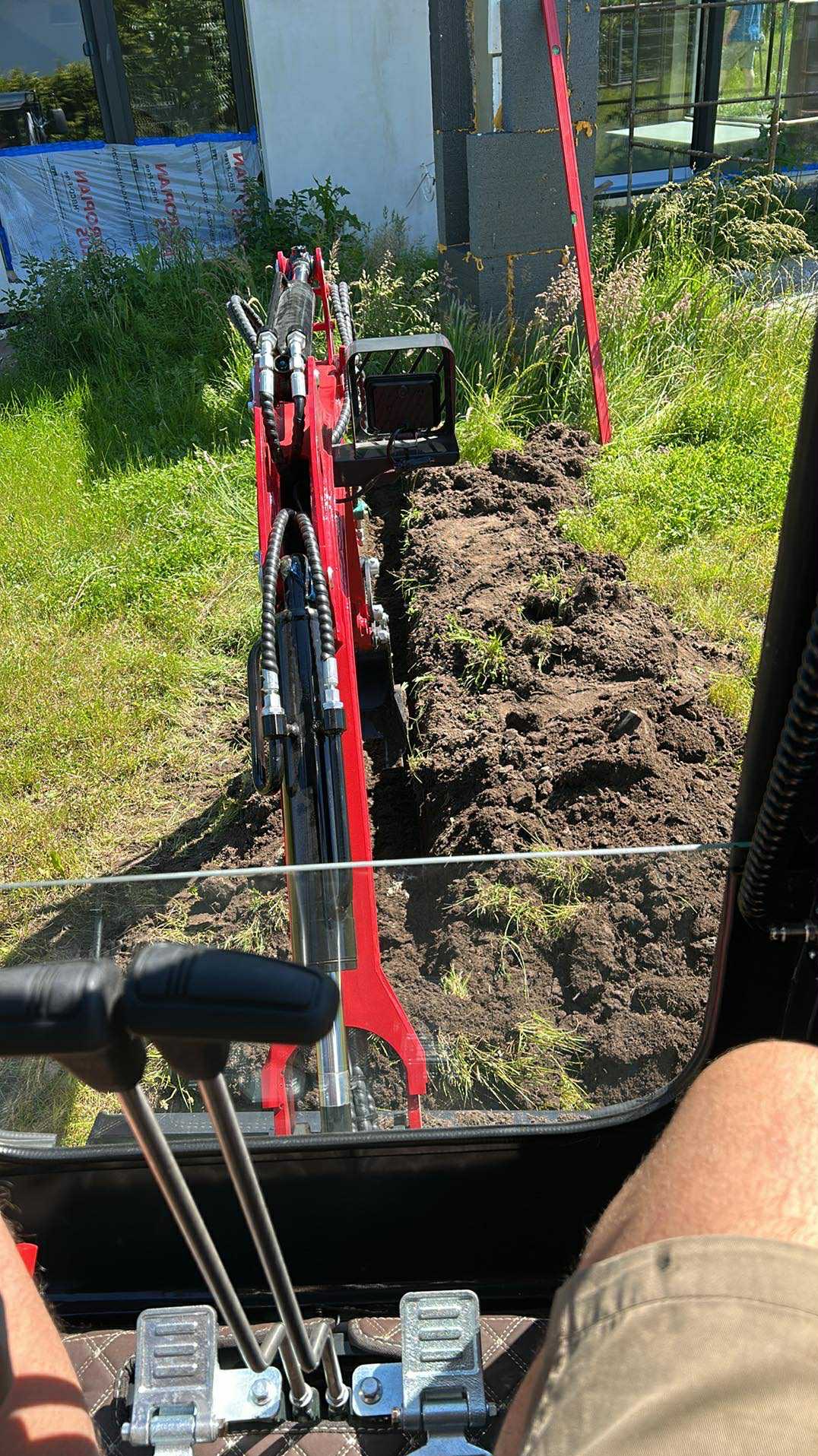 Czerwona mini koparka z perspektywy operatora, wykonująca wykop w gruncie. Widoczny budynek w tle i poziomica oparta o ścianę.