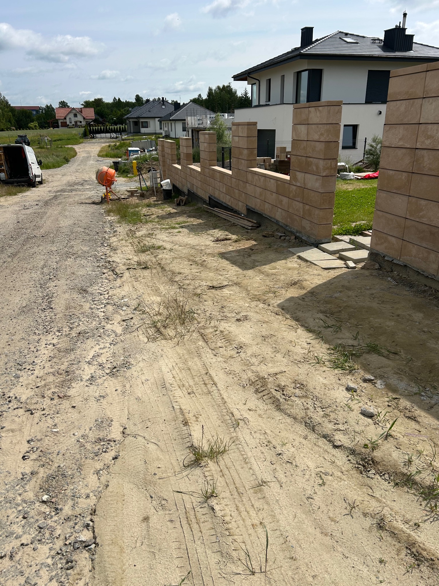 Montaż ogrodzenia z bloczków betonowych na posesji. Widoczna betoniarka, teren budowy i dom w tle. Elementy ogrodzenia w trakcie wznoszenia.