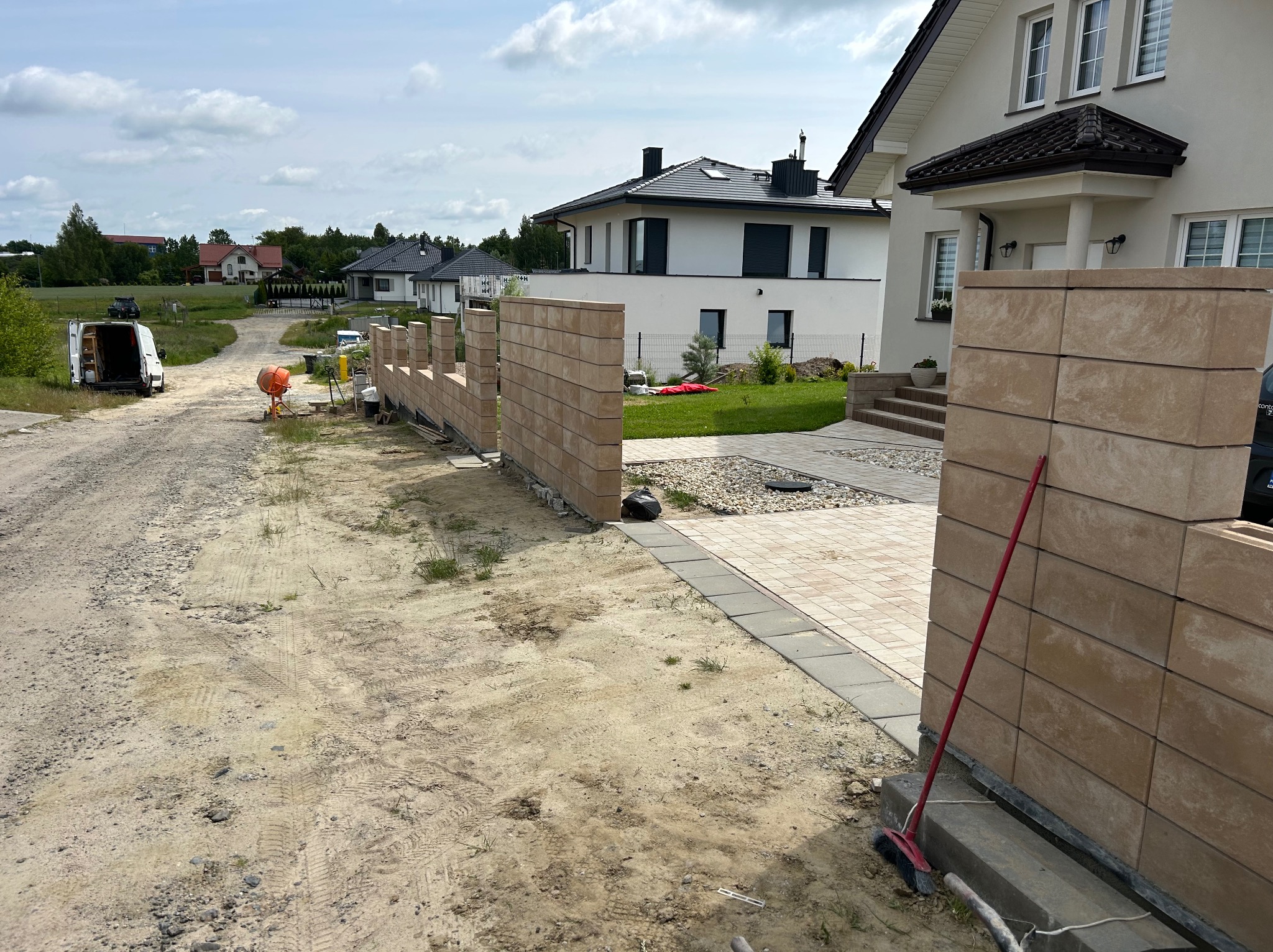 Budowa ogrodzenia betonowego modułowego na posesji. Widoczna betoniarka, częściowo ukończony mur i narzędzia. W tle domy jednorodzinne i zaparkowany samochód dostawczy.