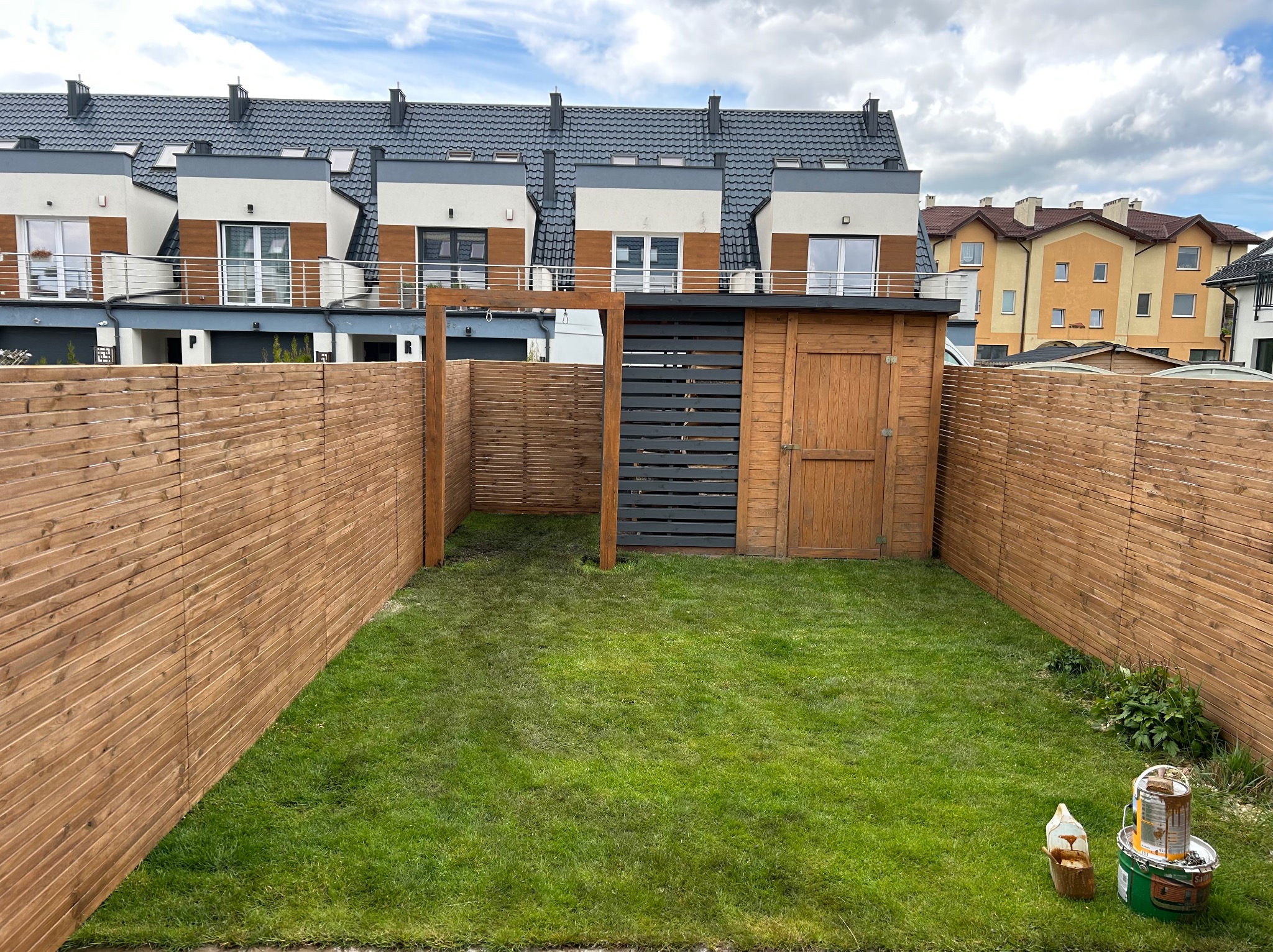 Świeżo założony trawnik otoczony drewnianym płotem lamelowym, z drewnianą furtką i szopą na narzędzia. W rogu puszki z farbą.