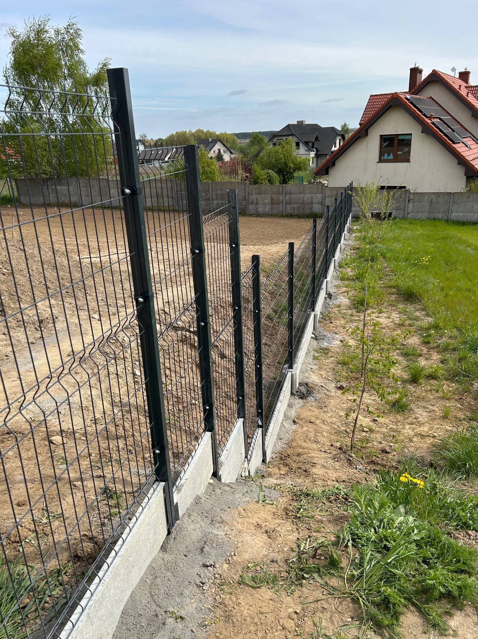 Nowoczesne ogrodzenie panelowe z podmurówką betonową, widok z boku na tle domów jednorodzinnych i zieleni. Zastosowano metalowe słupki i siatkę w kolorze grafitowym.