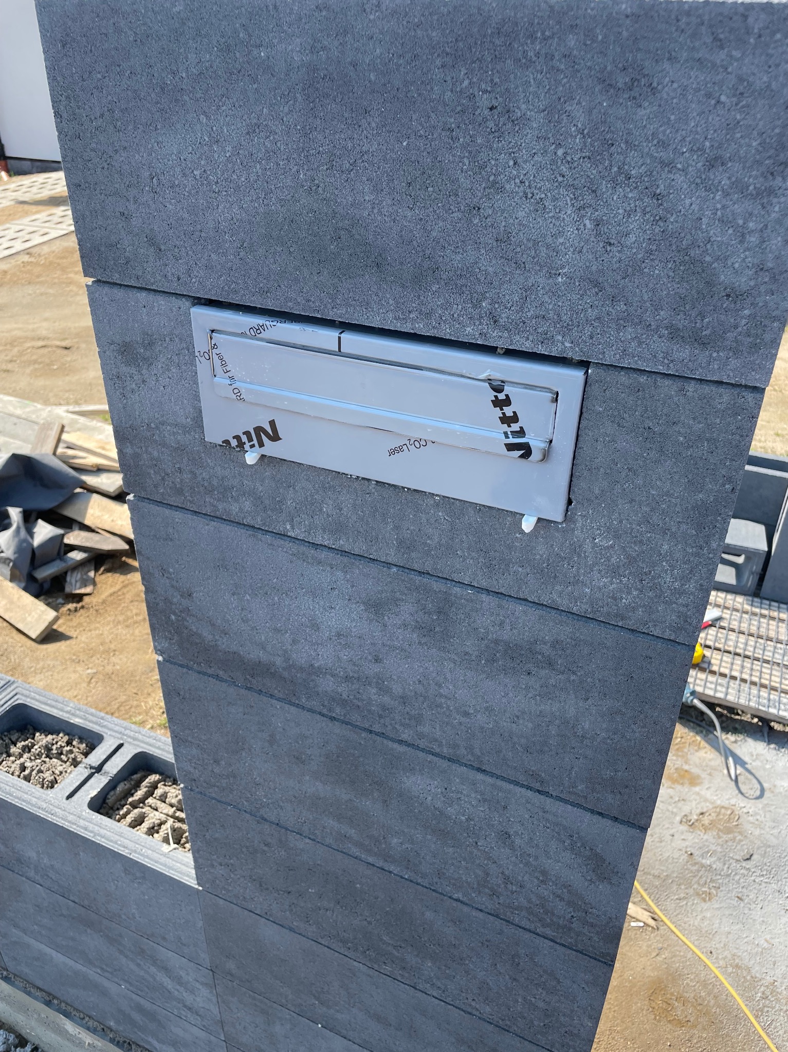 Szary, betonowy słupek ogrodzeniowy z zamontowaną, metalową skrzynką na listy. Widoczne ślady kleju montażowego i elementy konstrukcyjne ogrodzenia.