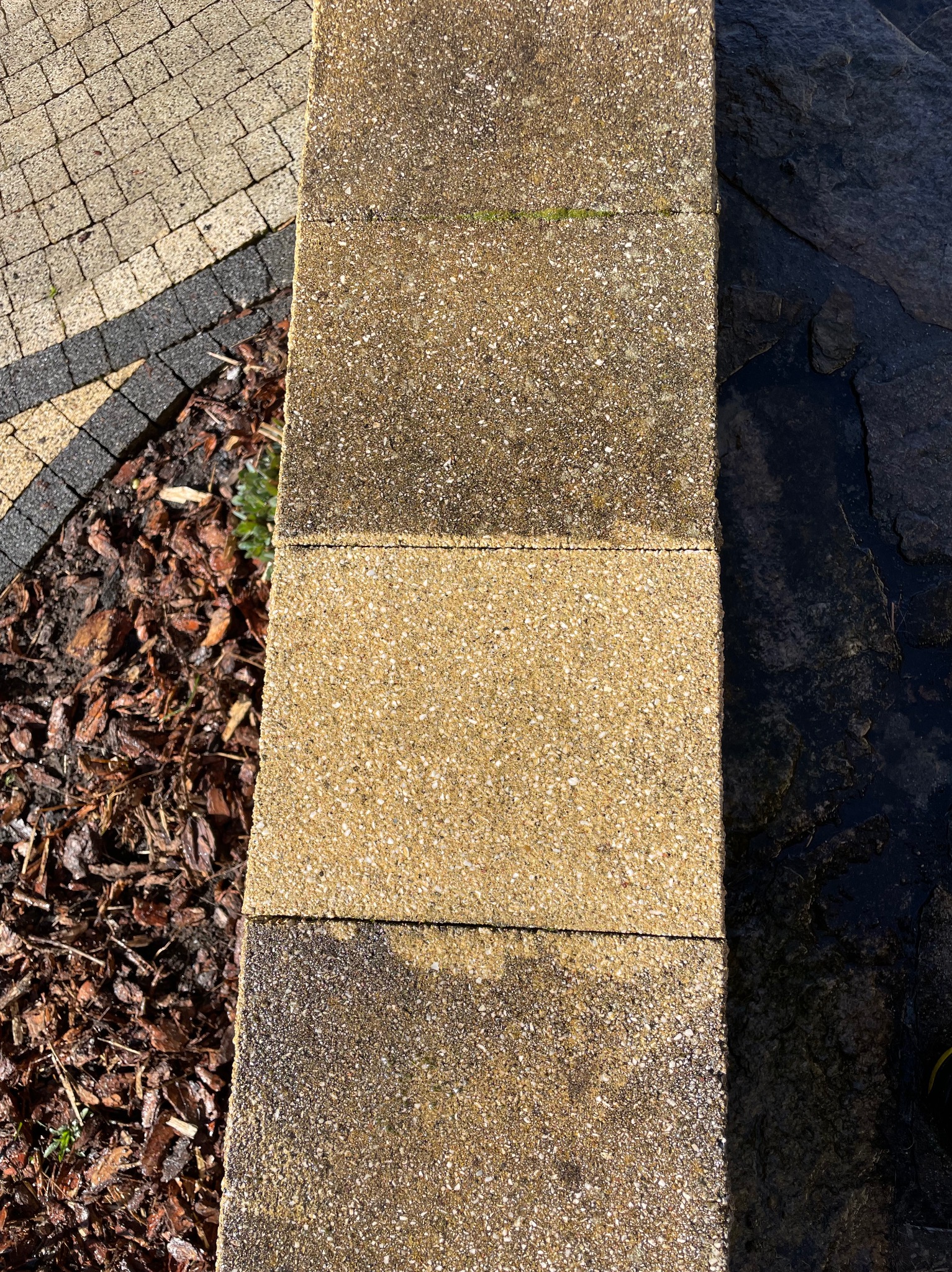 Ułożone, beżowe płyty chodnikowe z widoczną fakturą, obok kostki brukowej i kory drzewnej, tworzące ścieżkę w ogrodzie. Część płyt jest lekko wilgotna.