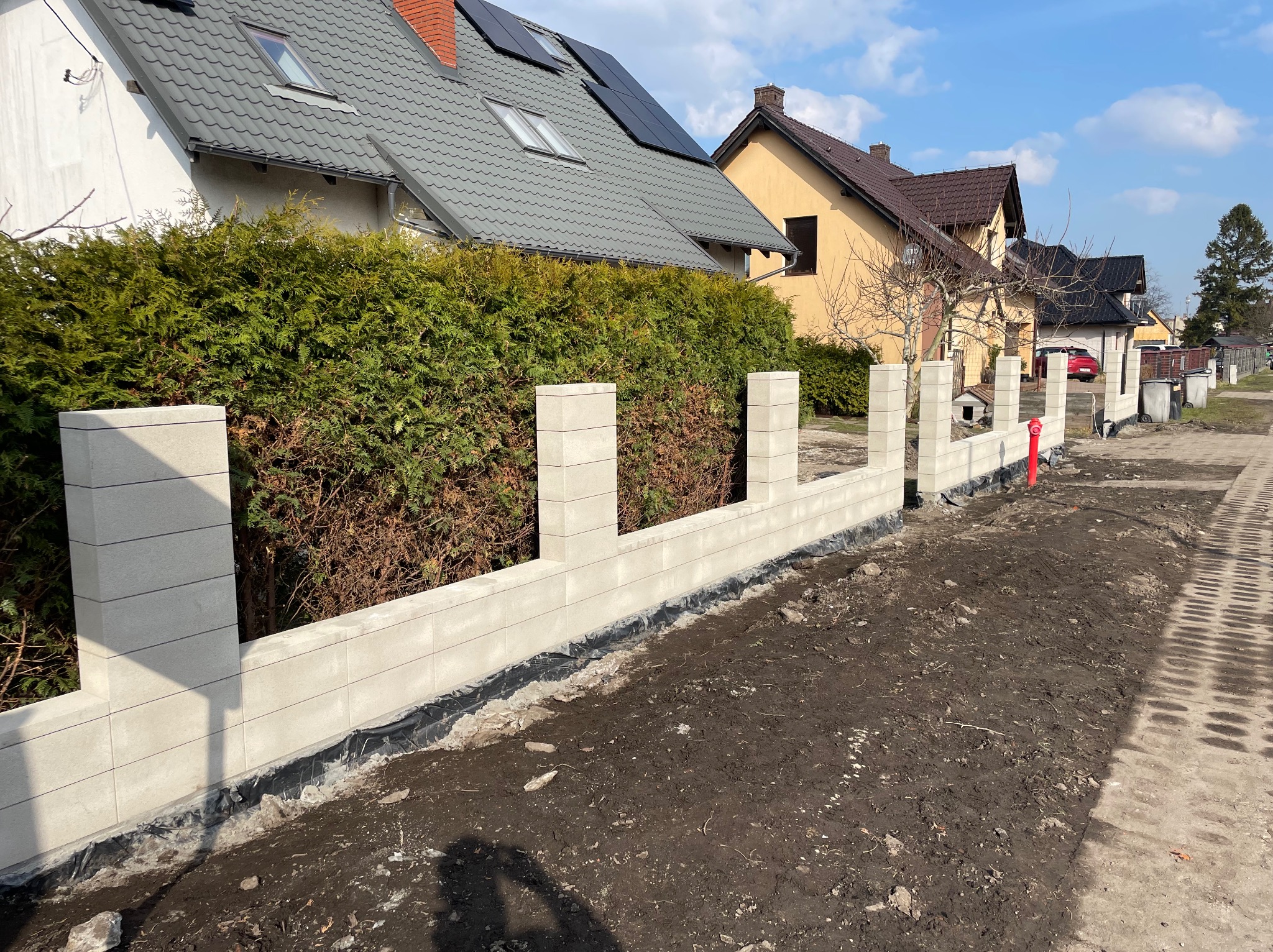 Wznoszenie ogrodzenia z prefabrykatów betonowych na posesji. Widoczne bloczki, fundament i częściowo zagospodarowany teren. W tle domy i błękitne niebo.