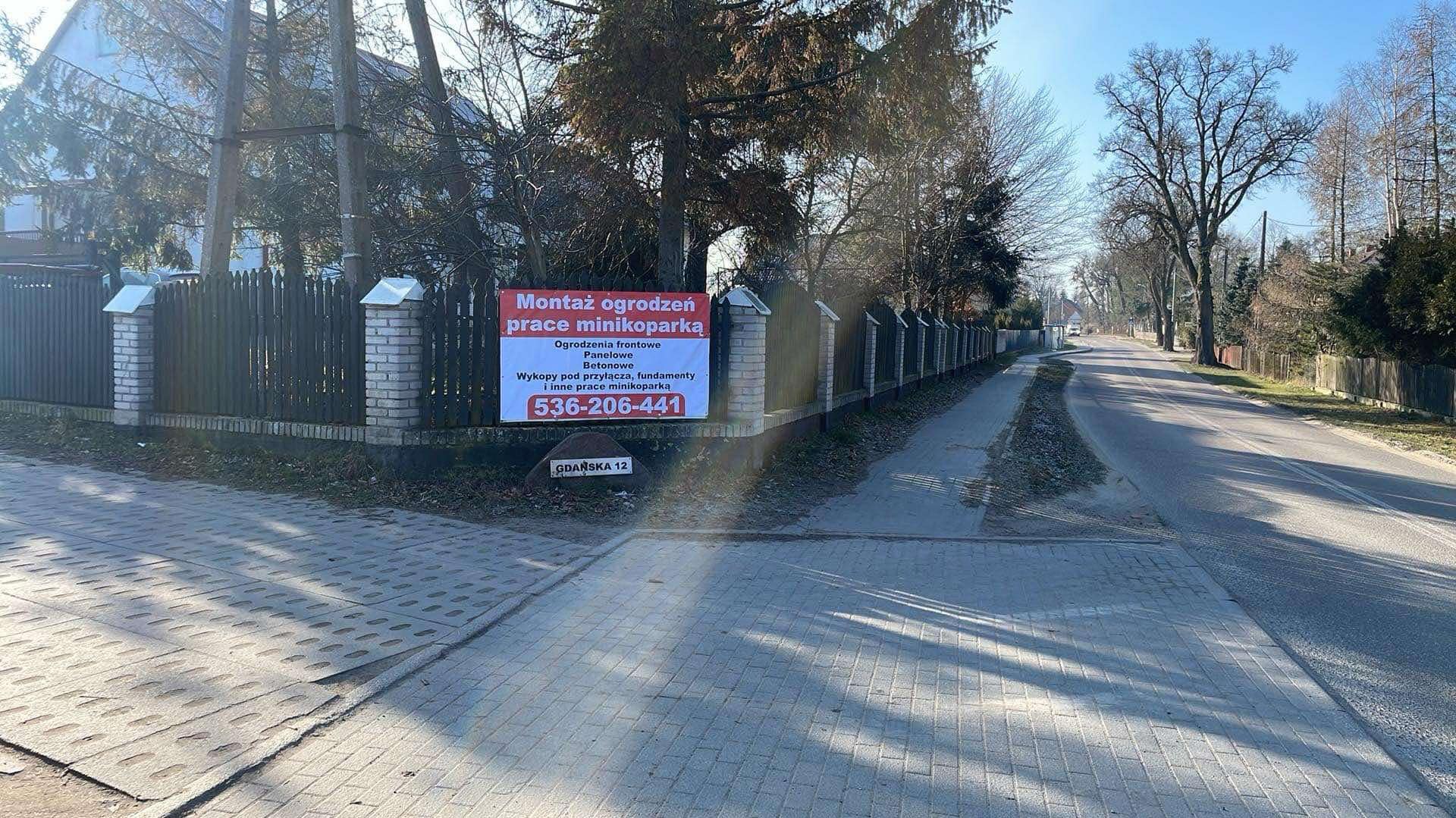 Ogrodzenie z drewnianymi przęsłami i betonowymi słupkami, z banerem reklamowym firmy montującej ogrodzenia i wykonującej prace minikoparką. Ujęcie z poziomu chodnika.