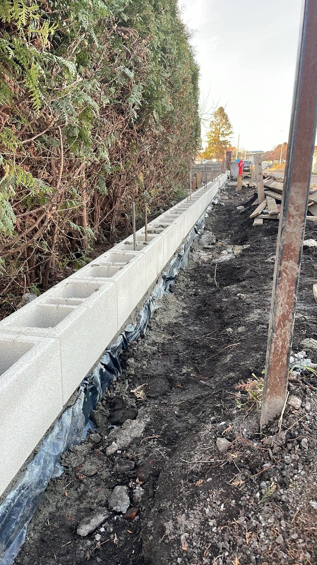 Wznoszenie fundamentu pod ogrodzenie z bloczków betonowych. Widoczne zbrojenie i folia izolacyjna. Tło: tuje i drzewa. Perspektywa z dołu, podkreślająca długość fundamentu.