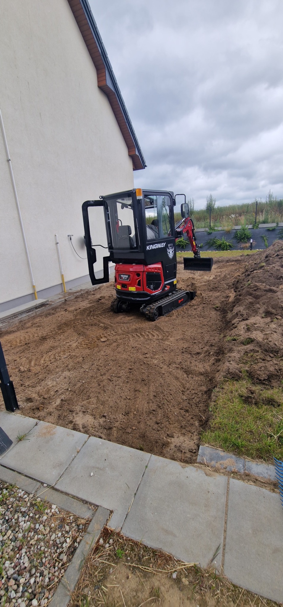 Czerwono-czarna mini koparka Kingway na przygotowanym terenie pod budowę ogrodzenia, widok z boku. W tle dom z jasną elewacją i fragment ogrodu.