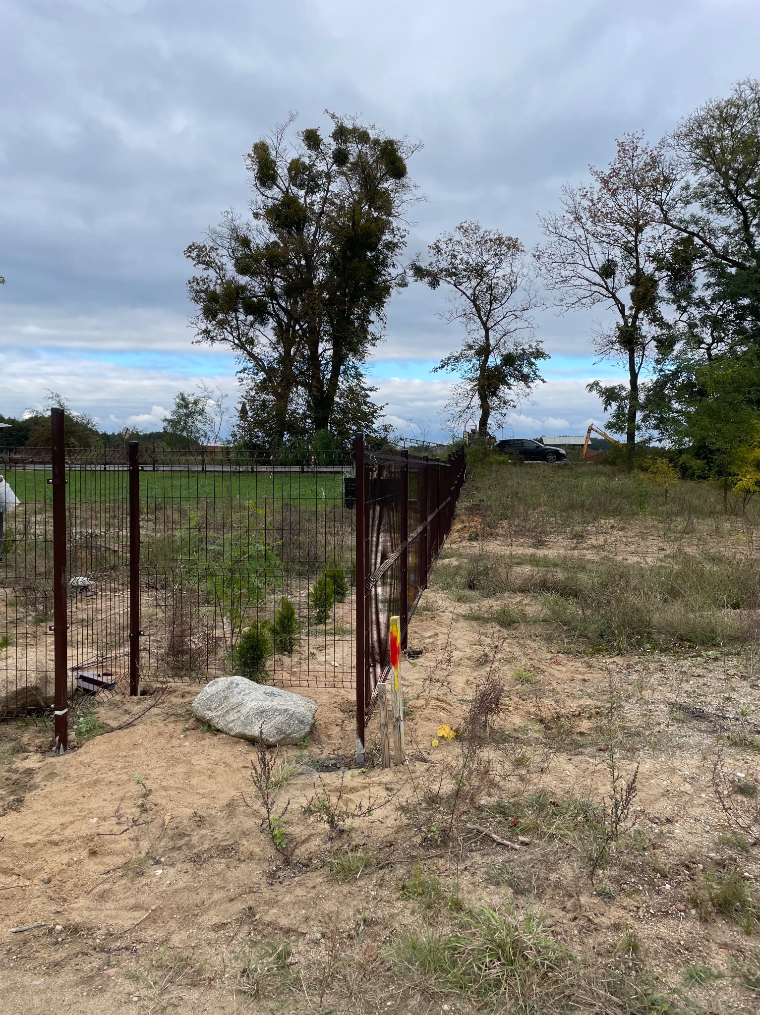Brązowe, metalowe ogrodzenie panelowe na nierównym, piaszczystym terenie. W tle drzewa i pochmurne niebo. Widoczny fragment koparki i zaparkowany samochód w oddali.