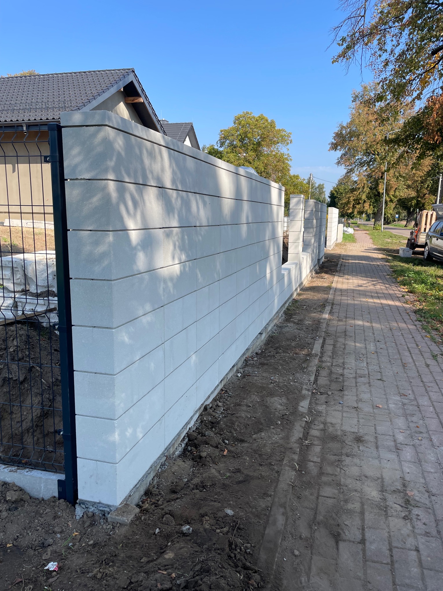 Nowoczesny, jasnoszary mur z prefabrykatów betonowych, połączony z metalową siatką ogrodzeniową. Widok na posesję i chodnik. Słoneczny dzień.