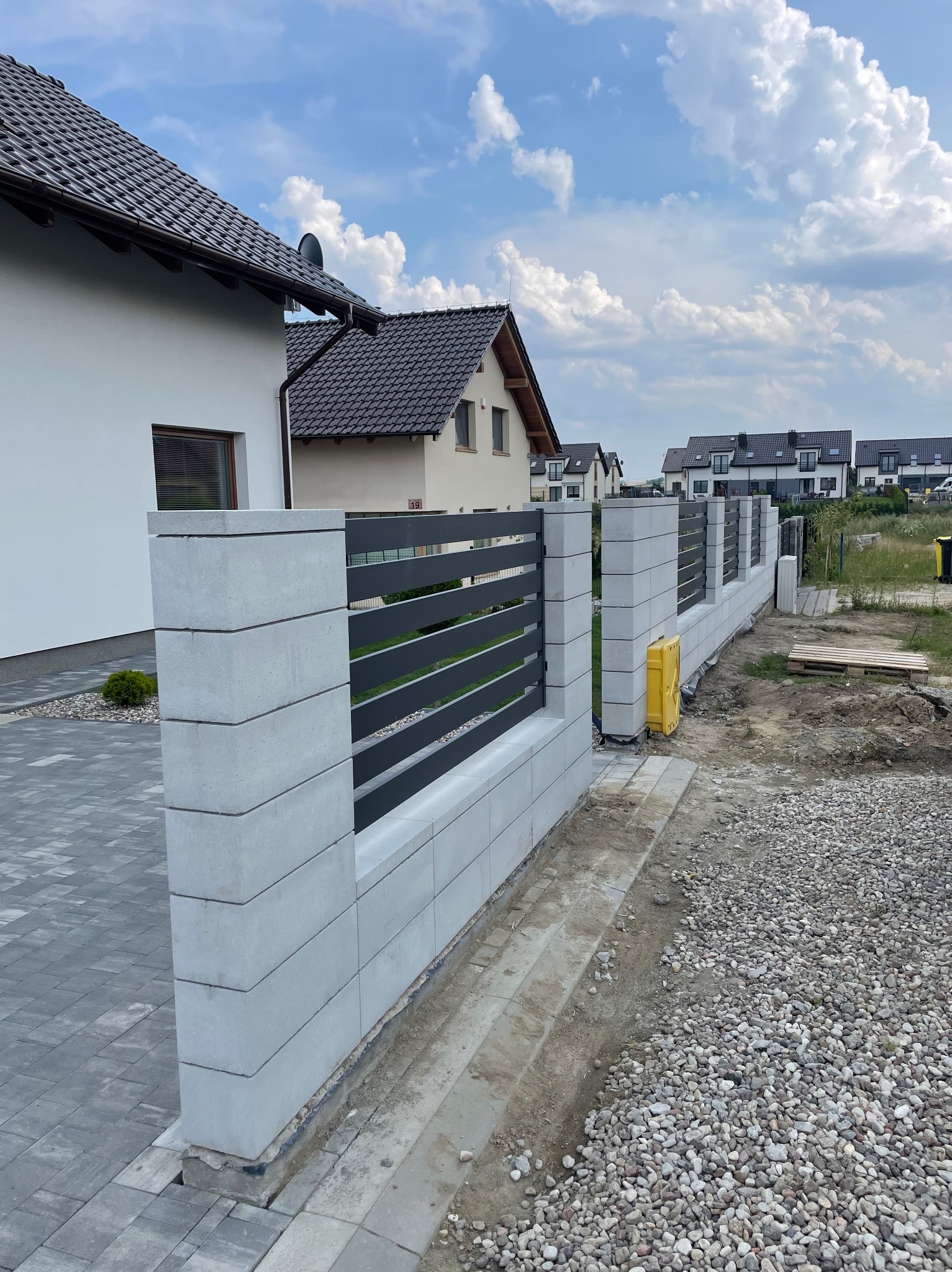 Nowoczesne ogrodzenie z betonowych słupków i metalowych przęseł w Kaczki. Widok na posesję i domy w tle, błękitne niebo. Minimalistyczny design, surowy beton, grafitowe elementy.