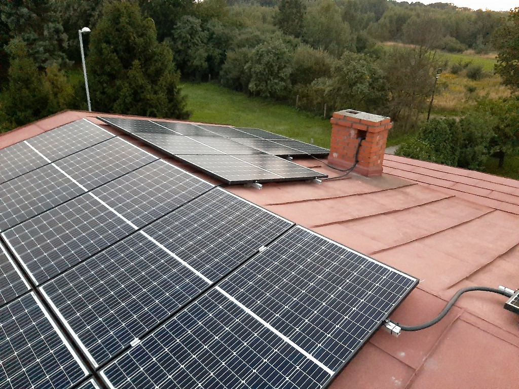 Instalacja paneli słonecznych na dachu z blachy na rąbek, widoczny komin z przewodami. Panele monokrystaliczne ułożone w rzędach, otoczenie zielone drzewa i trawa.