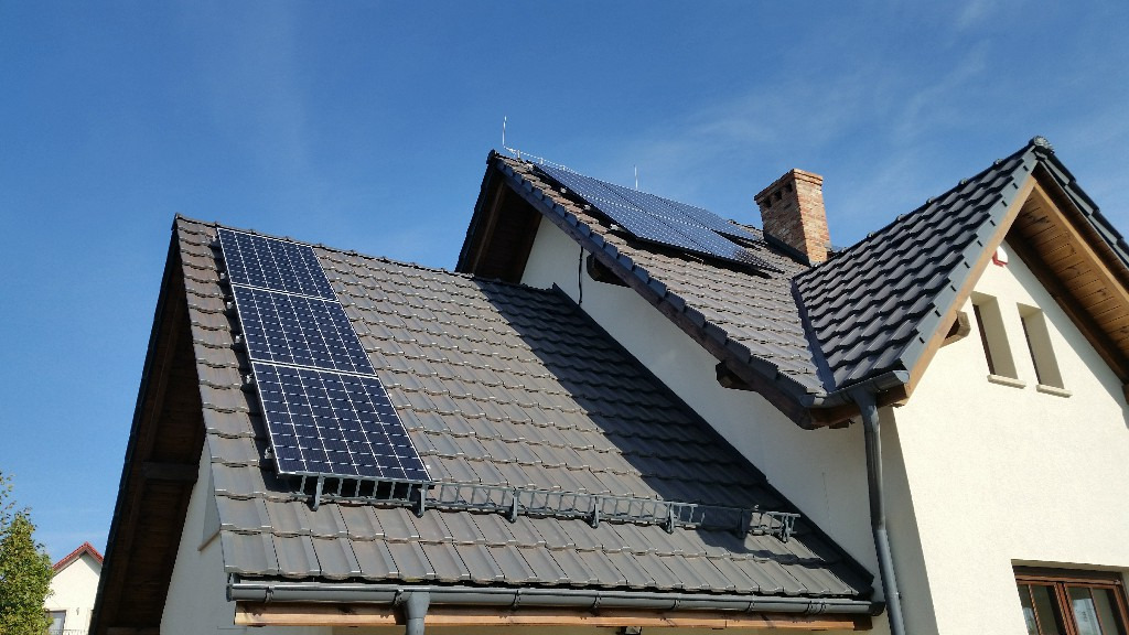 Panele słoneczne zamontowane na skośnym dachu domu jednorodzinnego z brązową dachówką, widoczne uchwyty montażowe paneli i komin w tle.
