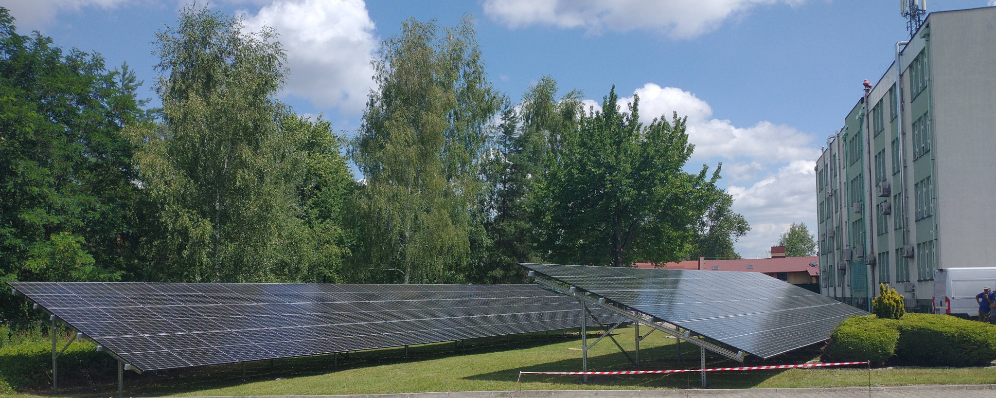 Instalacja fotowoltaiczna wolnostojąca na trawiastym terenie, z budynkiem w tle i pracownikiem na dachu, zabezpieczona taśmą ostrzegawczą.
