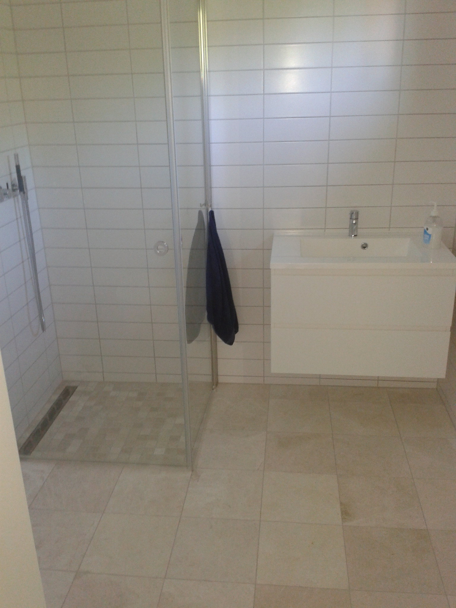 Nowoczesna łazienka z kabiną prysznicową ze szklanymi drzwiami, wiszącą białą szafką z umywalką i beżowymi płytkami na podłodze i ścianach.