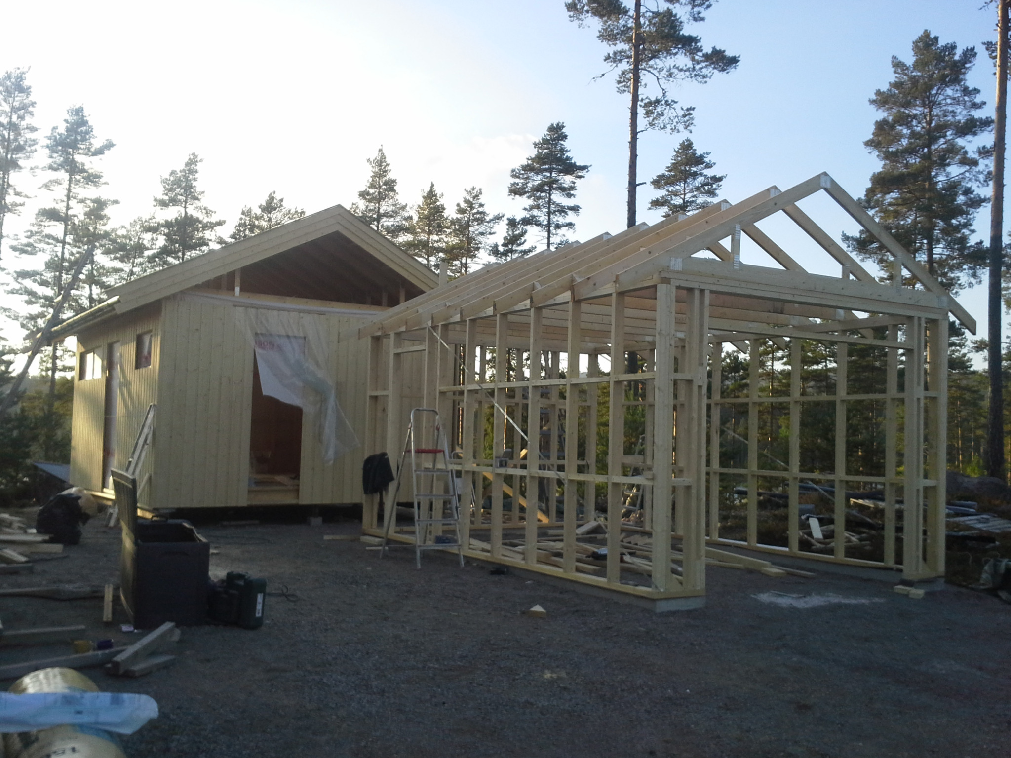 Budowa drewnianego domku letniskowego z dobudowaną wiatą garażową w lesie, widoczne elementy konstrukcyjne i drabina.