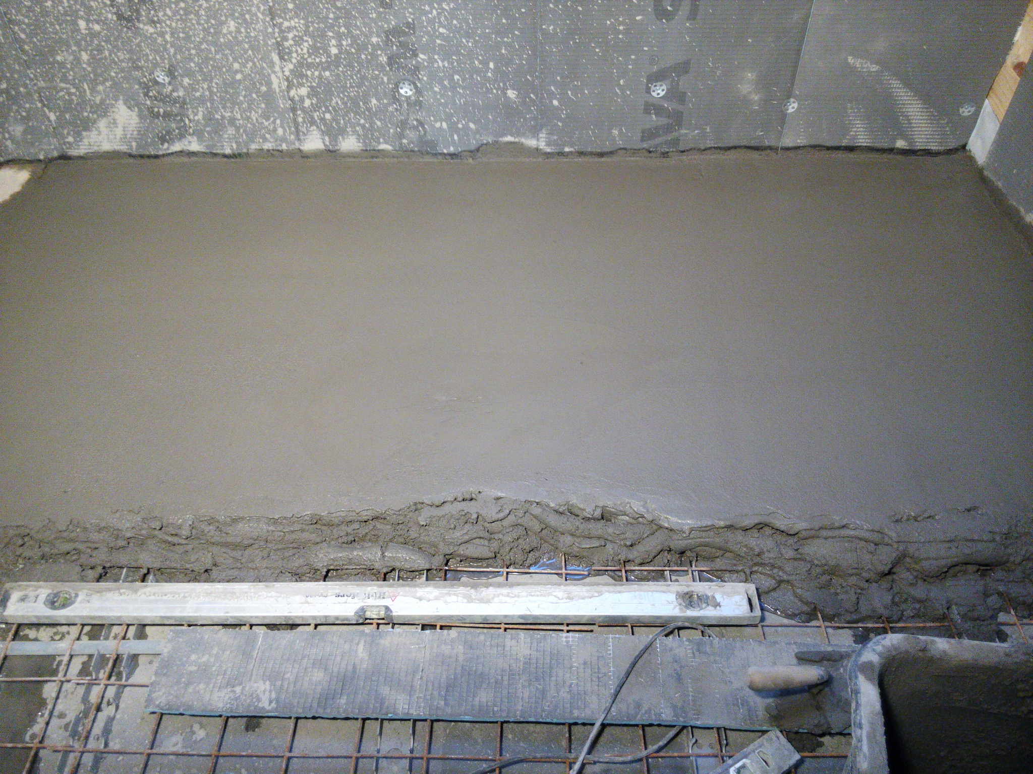 Wylewka betonowa na siatce zbrojeniowej, wyrównywana łatą, widoczne fragmenty ściany z szarą membraną izolacyjną.
