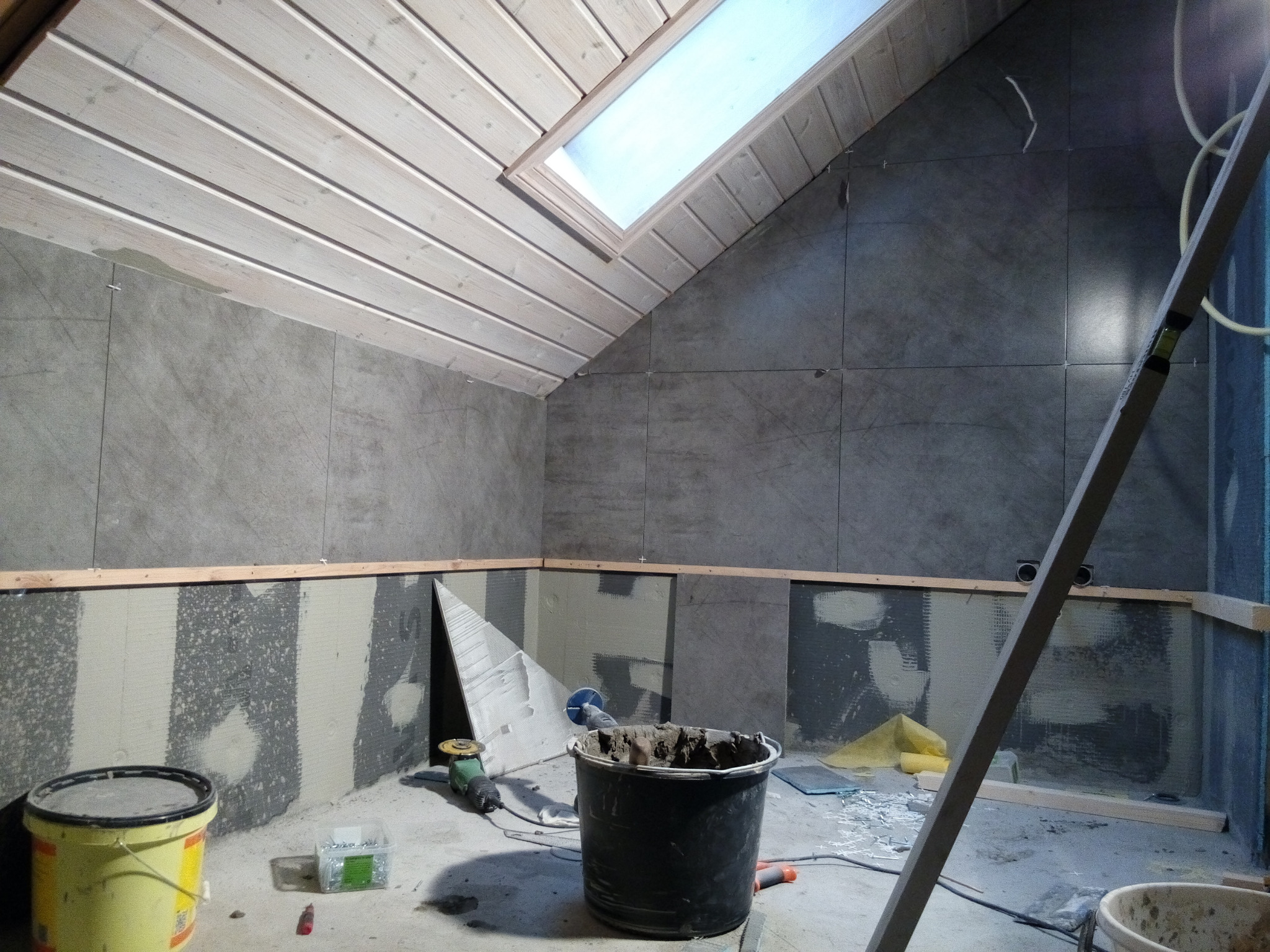 Montaż szarych płytek w łazience na poddaszu ze skośnym sufitem, widoczne narzędzia, wiadro z zaprawą, poziomica i okno dachowe.