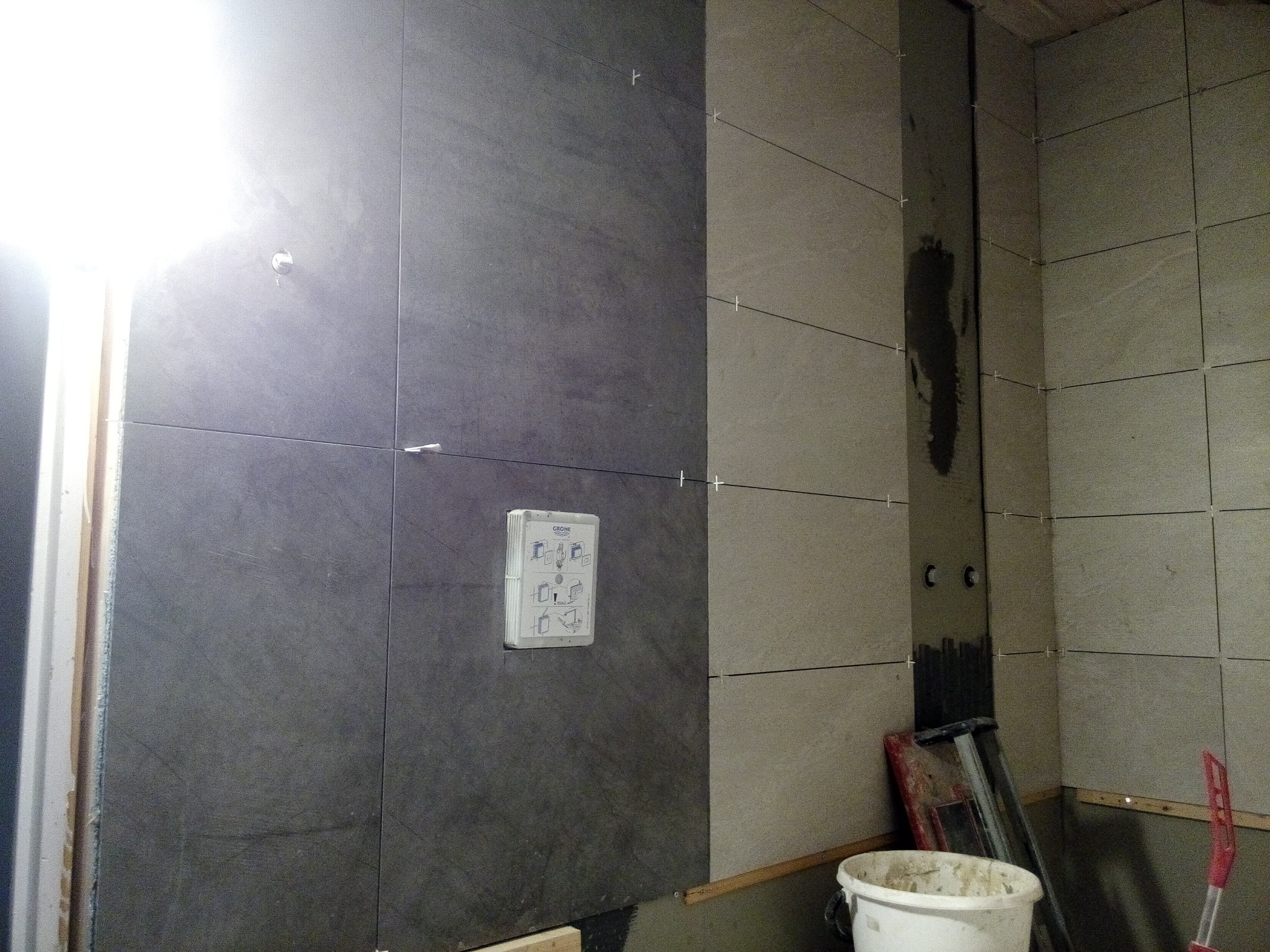 Wnętrze łazienki w trakcie układania płytek ceramicznych w dwóch odcieniach szarości, widoczne krzyżyki dystansowe, instalacja podtynkowa Grohe oraz narzędzia pracy.